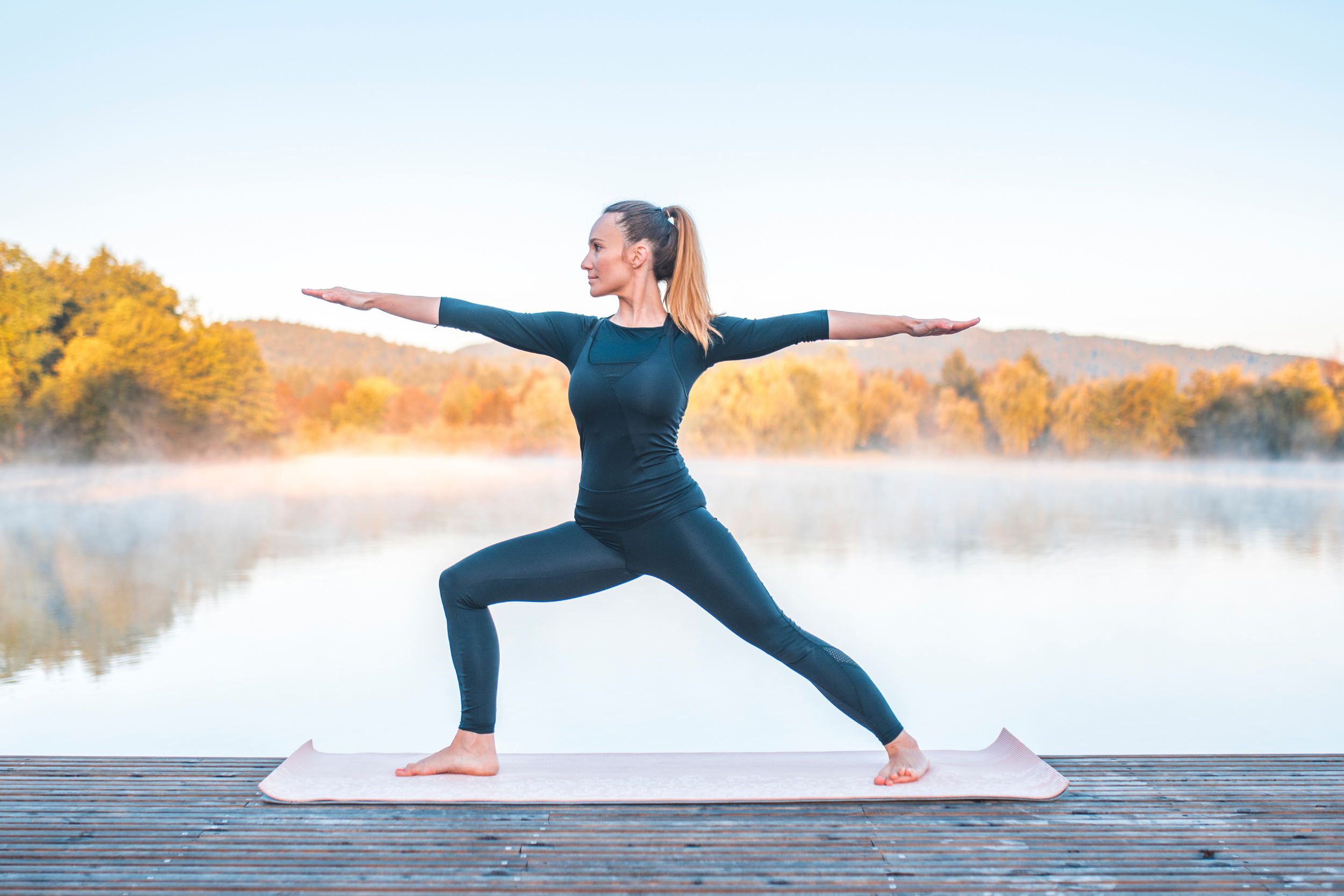 woman wearing black top and black leggins performing a warrior 2 on a deck with a lake and trees in the background