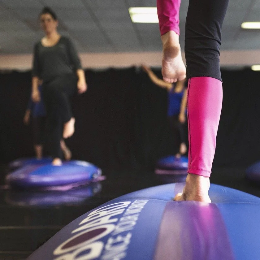 close up on a barefoot instructor performing a balance pose on a balance board called B-BOARD® flipped over with some participants doing the same in the background, in a fitness room