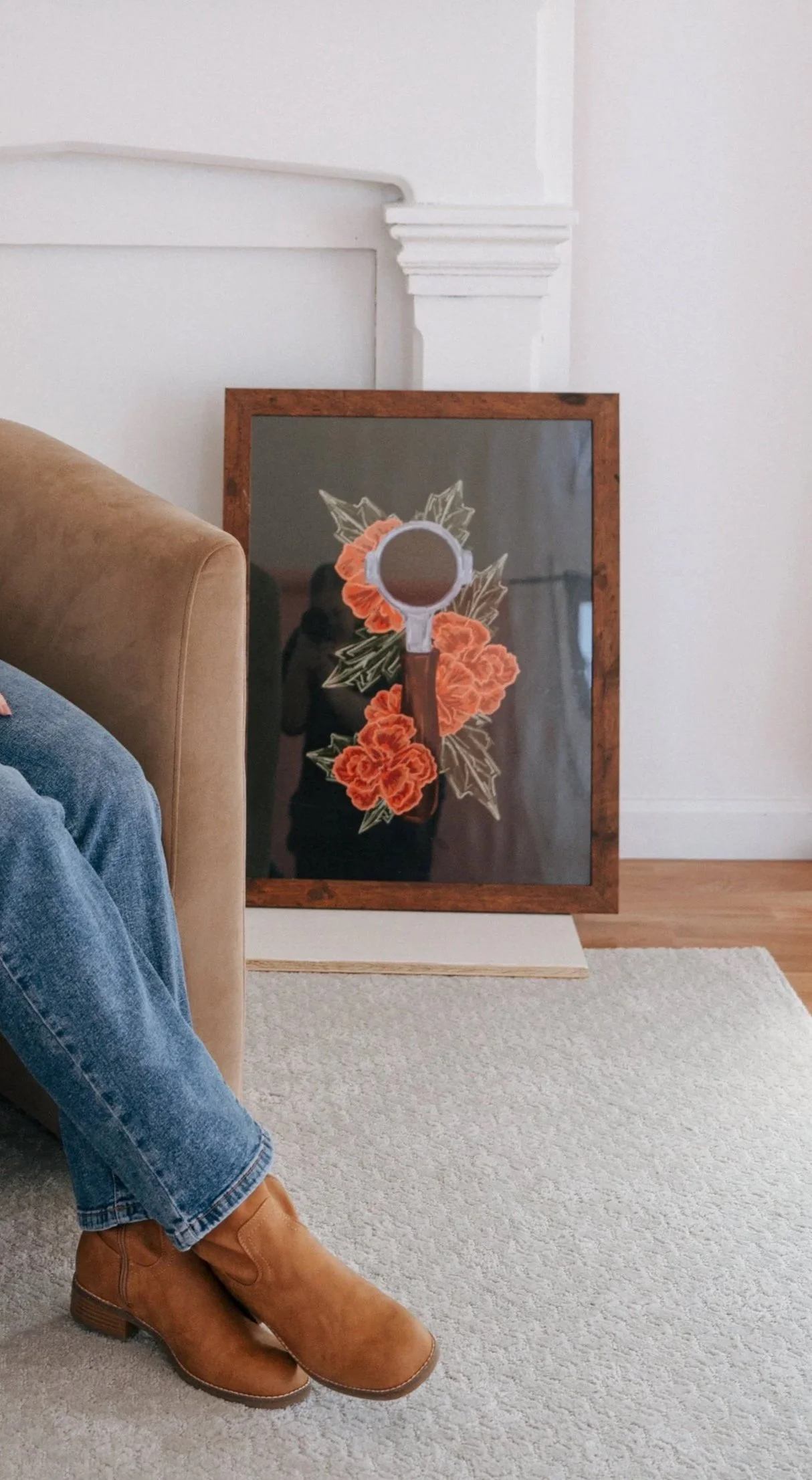 A mirror leaning against the wall with a painted rose and foliage design, partially obscured by a beige sofa and a person's leg wearing jeans and brown boots, on a white rug in a room with hardwood floor.