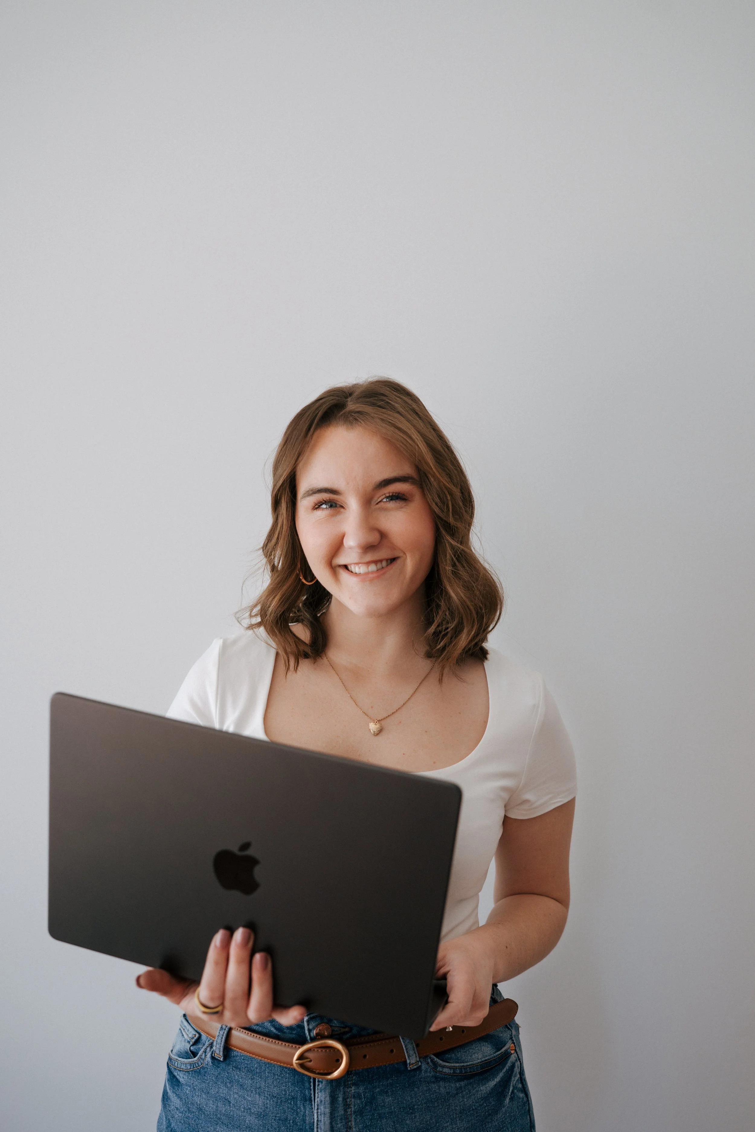 A young woman with shoulder-length wavy brown hair, wearing a white T-shirt and blue jeans, holding a closed MacBook laptop and smiling at the camera against a plain white wall.