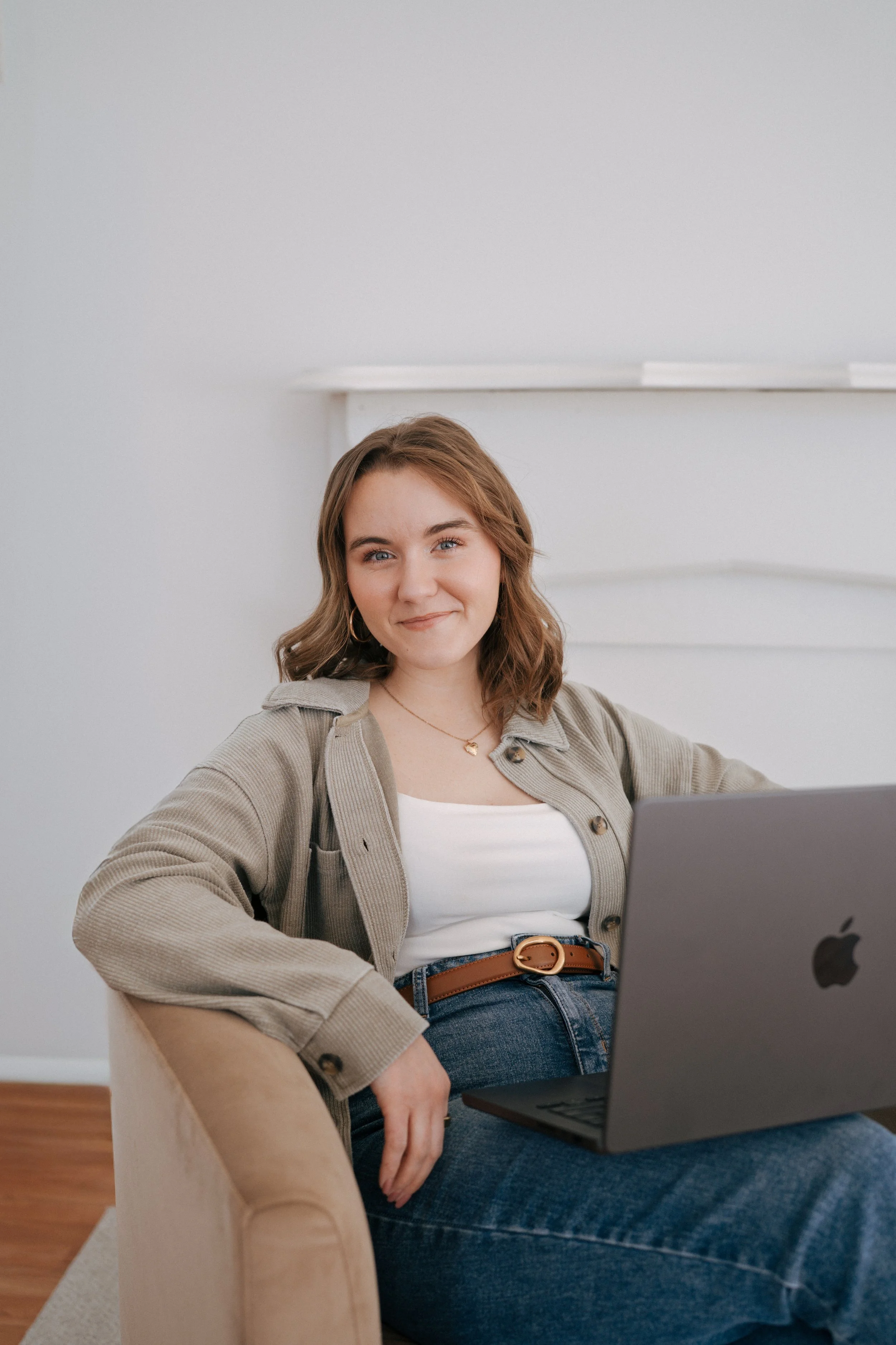 A young woman with shoulder-length wavy hair, wearing a beige jacket over a white top, sitting on a beige couch with a MacBook on her lap, smiling at the camera in a bright, minimalist room.