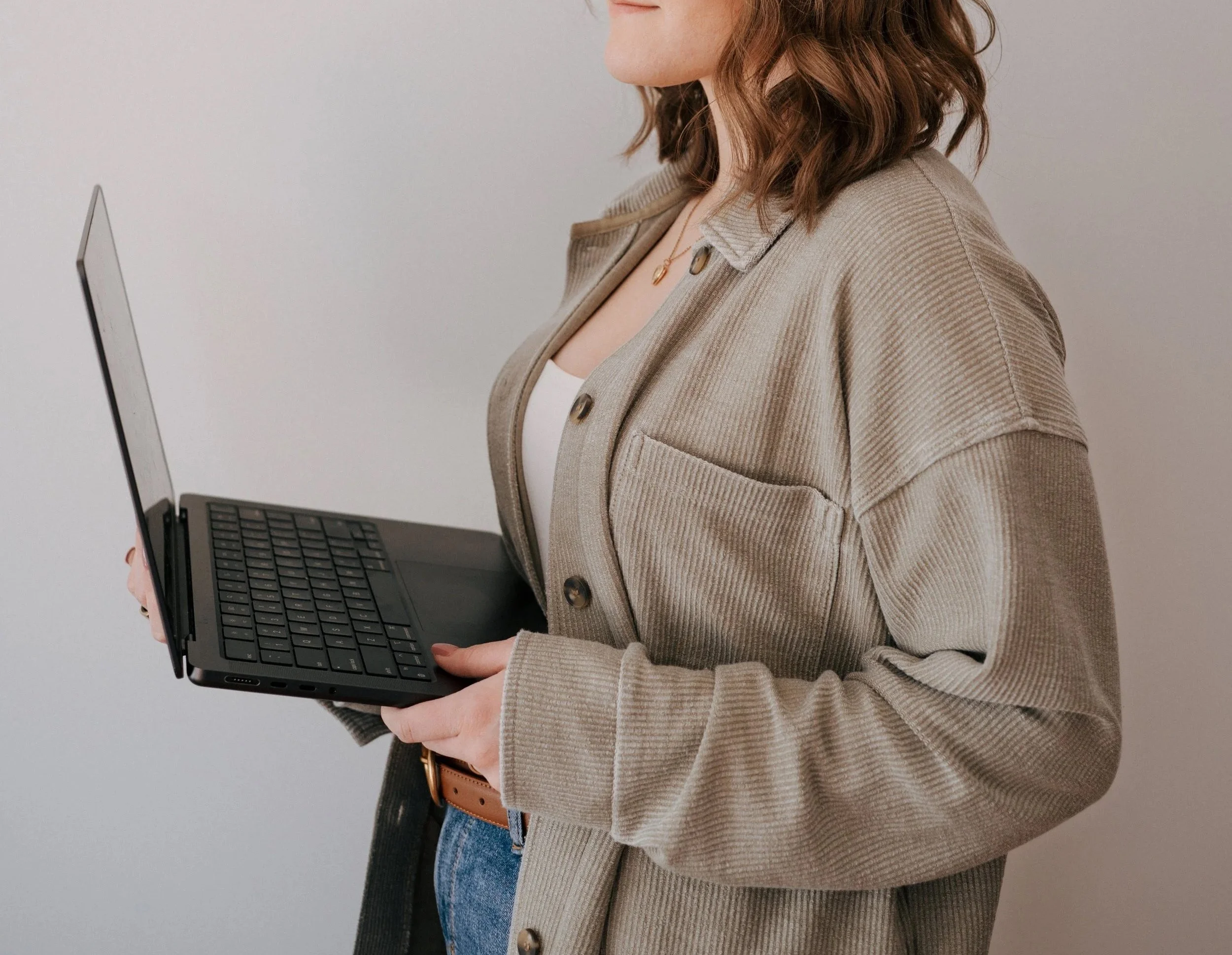 A woman with wavy brown hair holding a black laptop in her left hand and wearing a beige jacket, white top, blue jeans, and a brown belt.