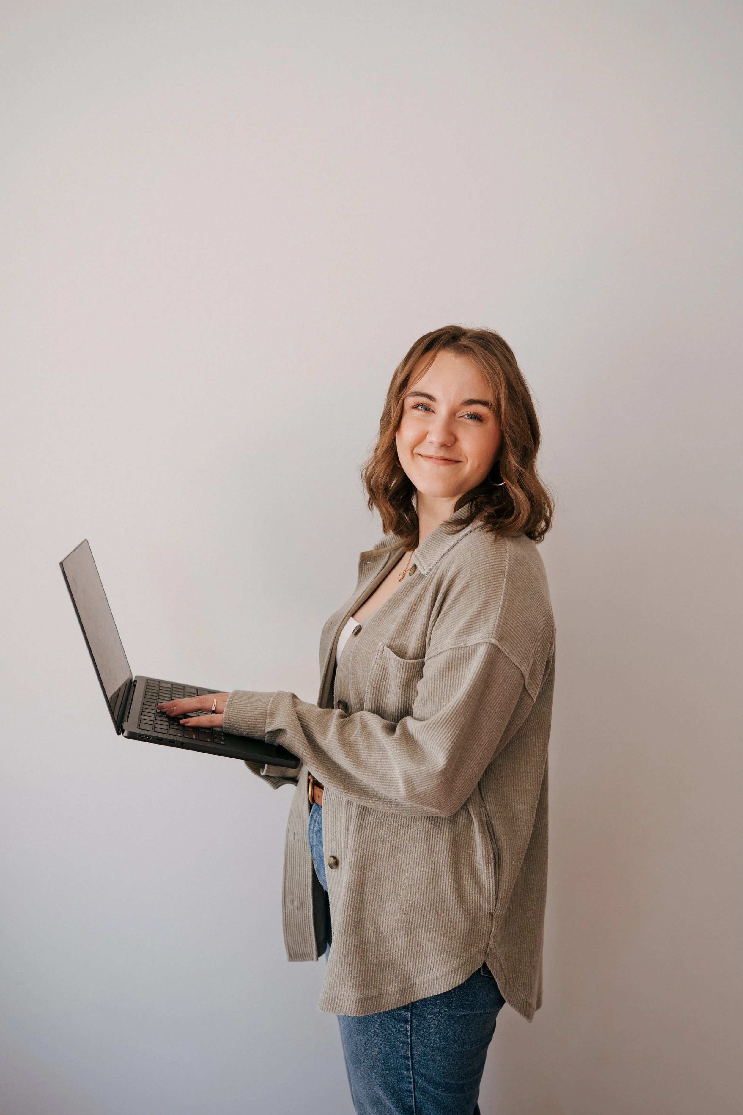 A young woman with shoulder-length brown hair, wearing a beige jacket and blue jeans, holding a laptop and smiling at the camera.
