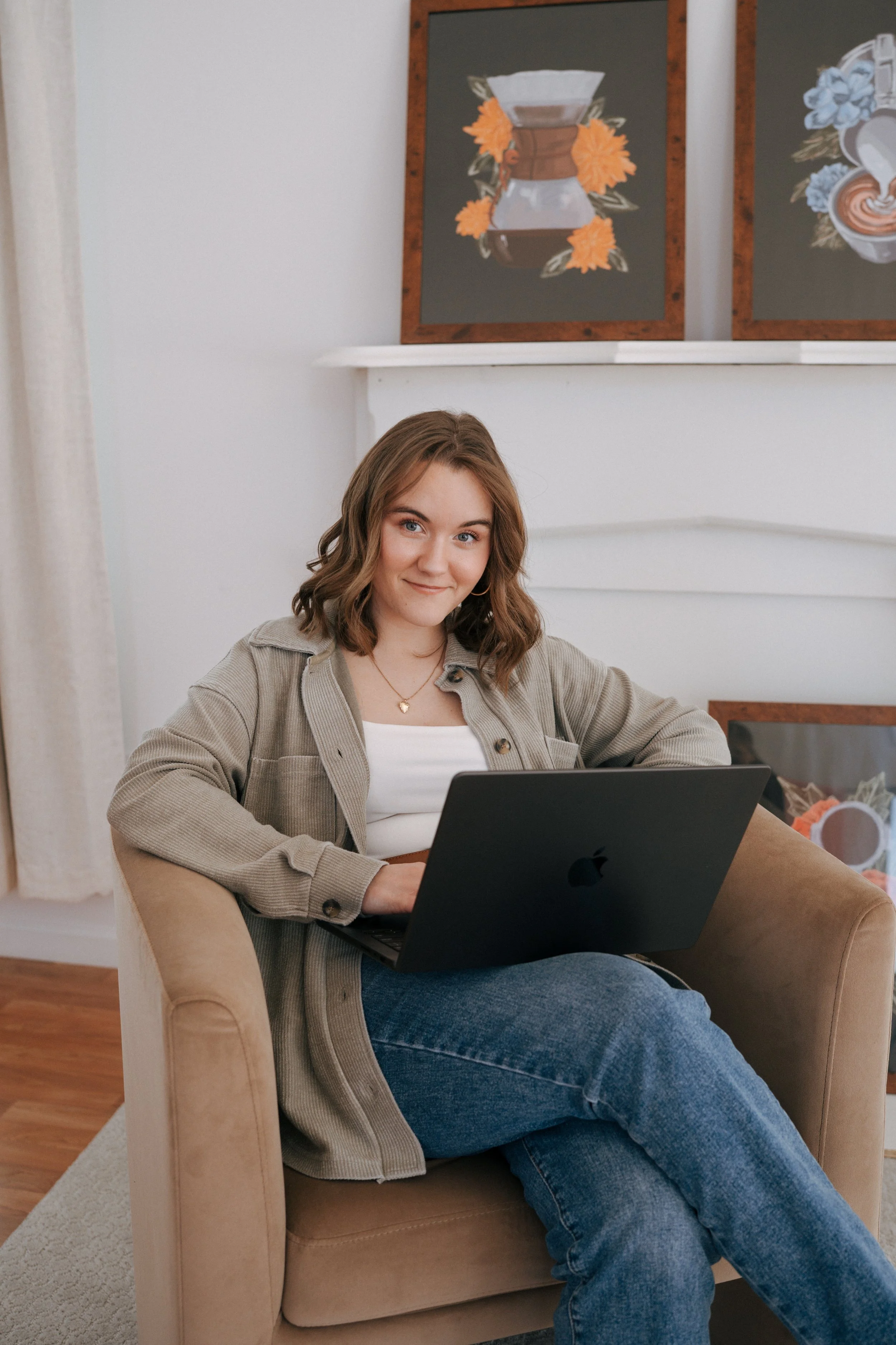 A young woman with shoulder-length brown hair, wearing a beige jacket and white top, sitting on a tan armchair while using a black MacBook laptop in a cozy living room.  Artwork on the wall behind her shows a coffee pot surrounded by orange and blue flowers.