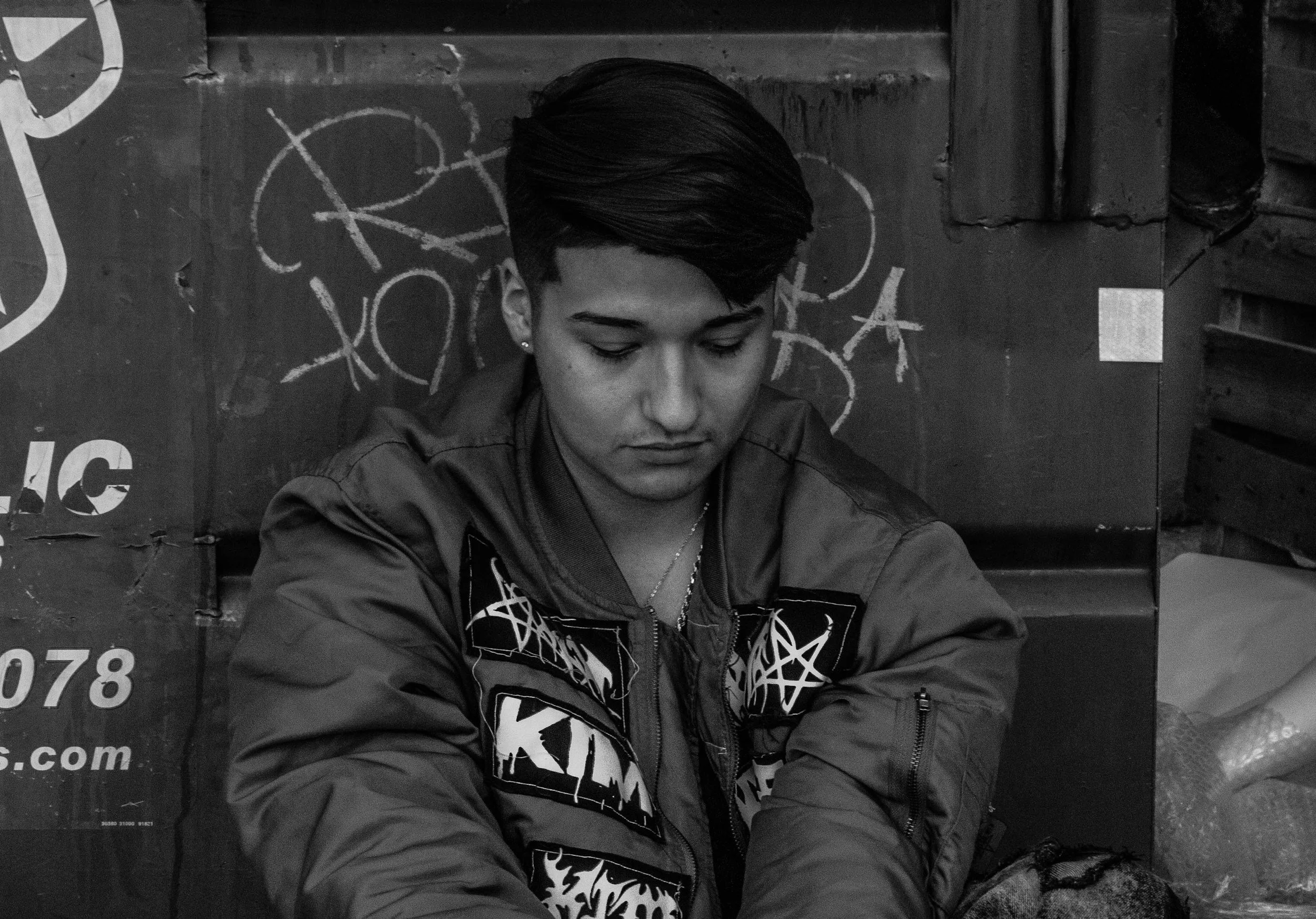 A young person with short dark hair looking down, sitting against a graffiti-covered wall, wearing a patterned jacket.
