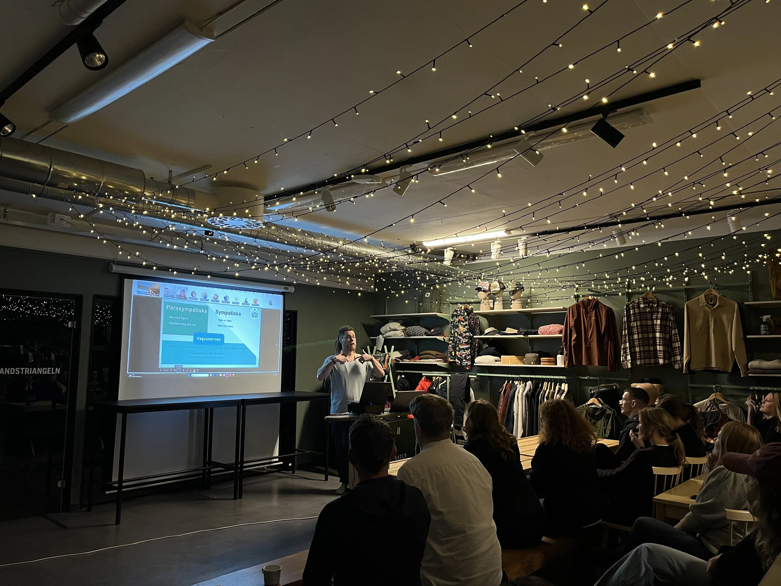 En person håller en företags presentation om breathwork medveten andning inför en grupp människor i ett rum dekorerat med string lights i taket. På skärmen visas en presentation med diagram och text