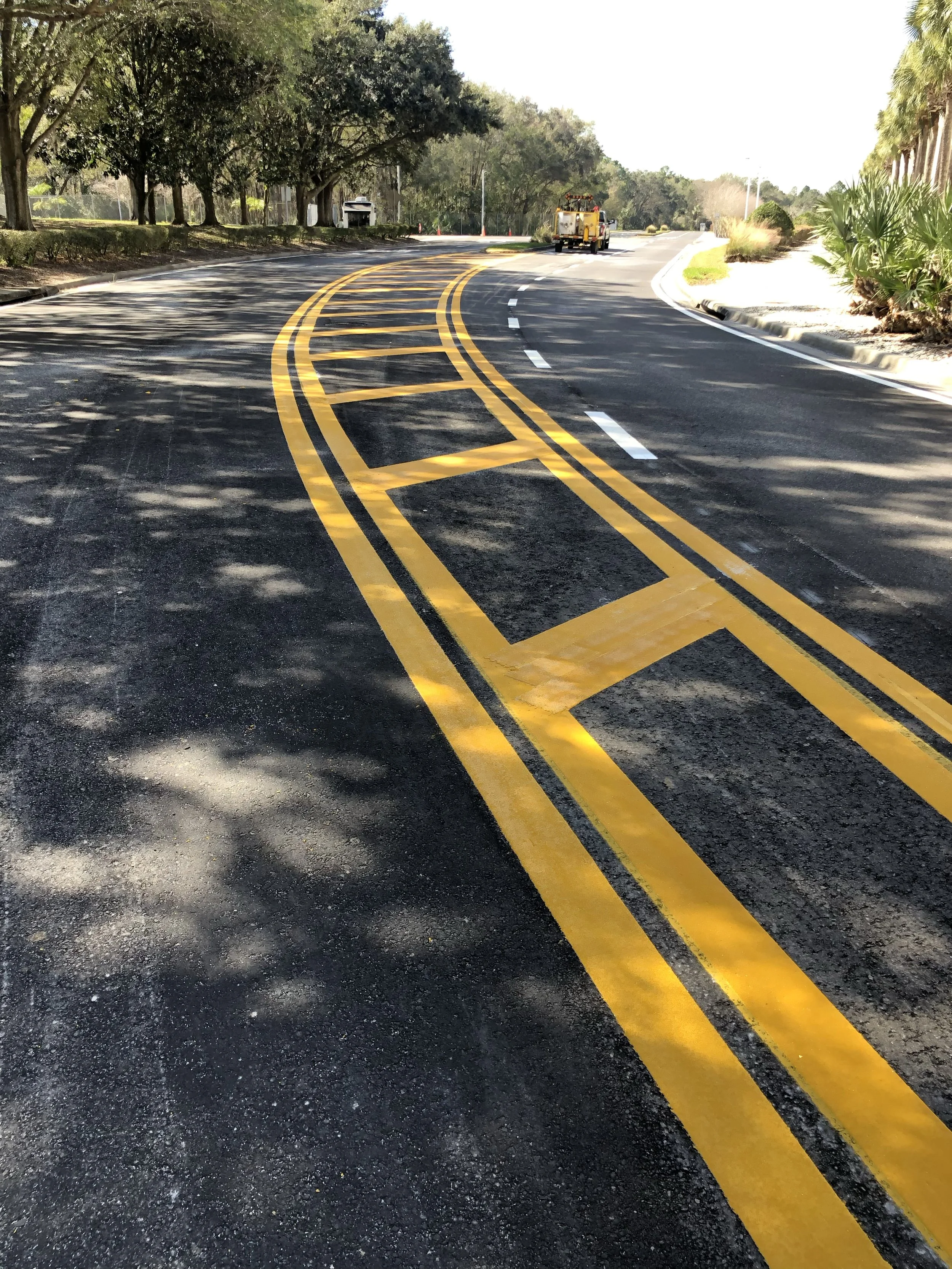 A cleanly paved FDOT road with bright yellow and white road markings, curving through a lush green area with trees and bushes.