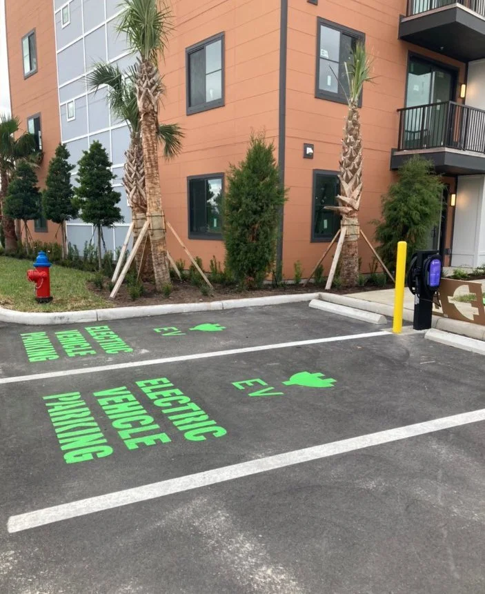 Electric vehicle charging stations with green markings on asphalt, labeled 'ELECTRIC VEHICLE' and 'SV,' with a fire hydrant, trees, and a multi-story building in the background.