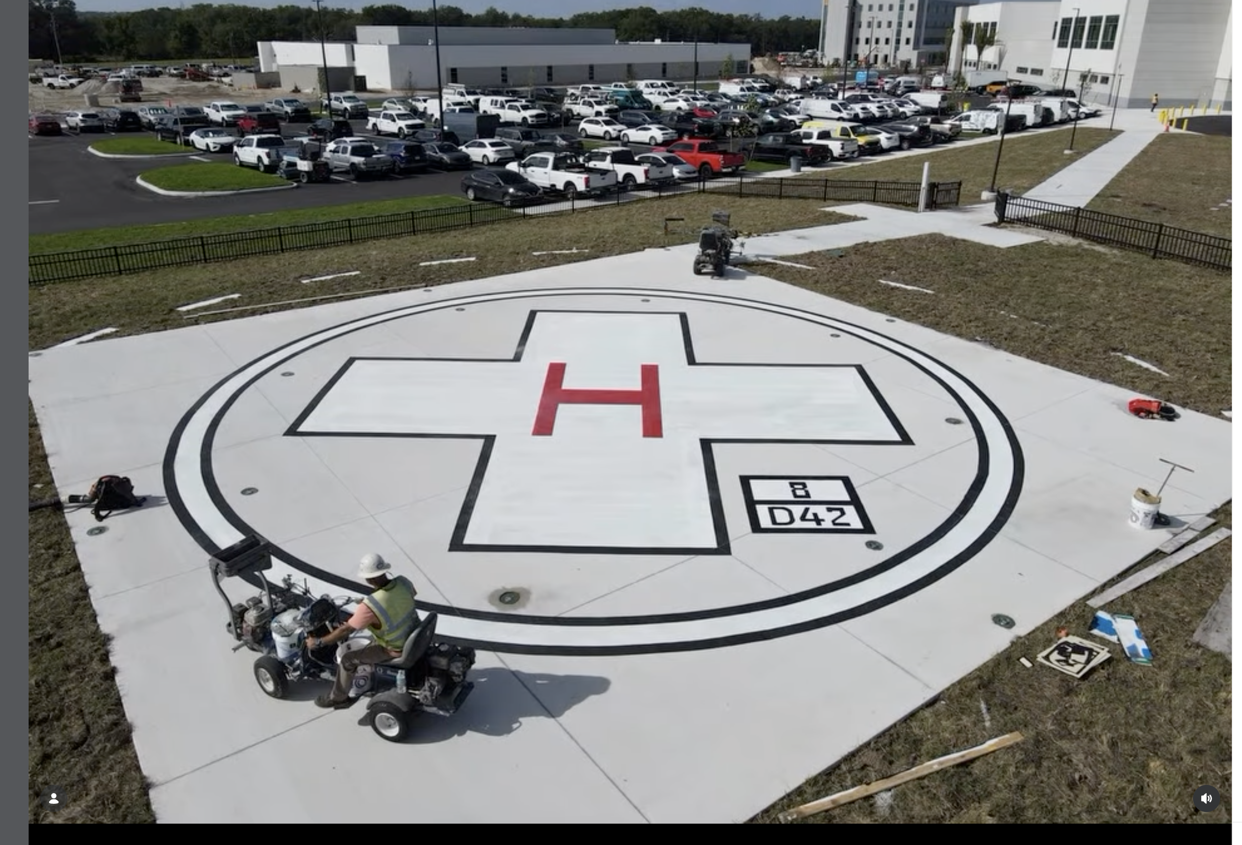 Workers are painting a helicopter landing pad with a large red H and black outlines. The area is outdoors, with a grassy area, a black fence, and a parking lot filled with white and black vehicles in the background. Equipment and tools are visible around the workers.