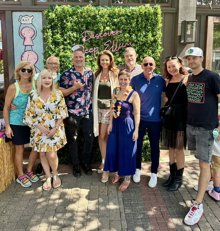 Group of ten people posing in front of a green hedge with a pink sign that says "Discover Deep Ellum". They are smiling and dressed casually, some wearing sunglasses and colorful outfits.