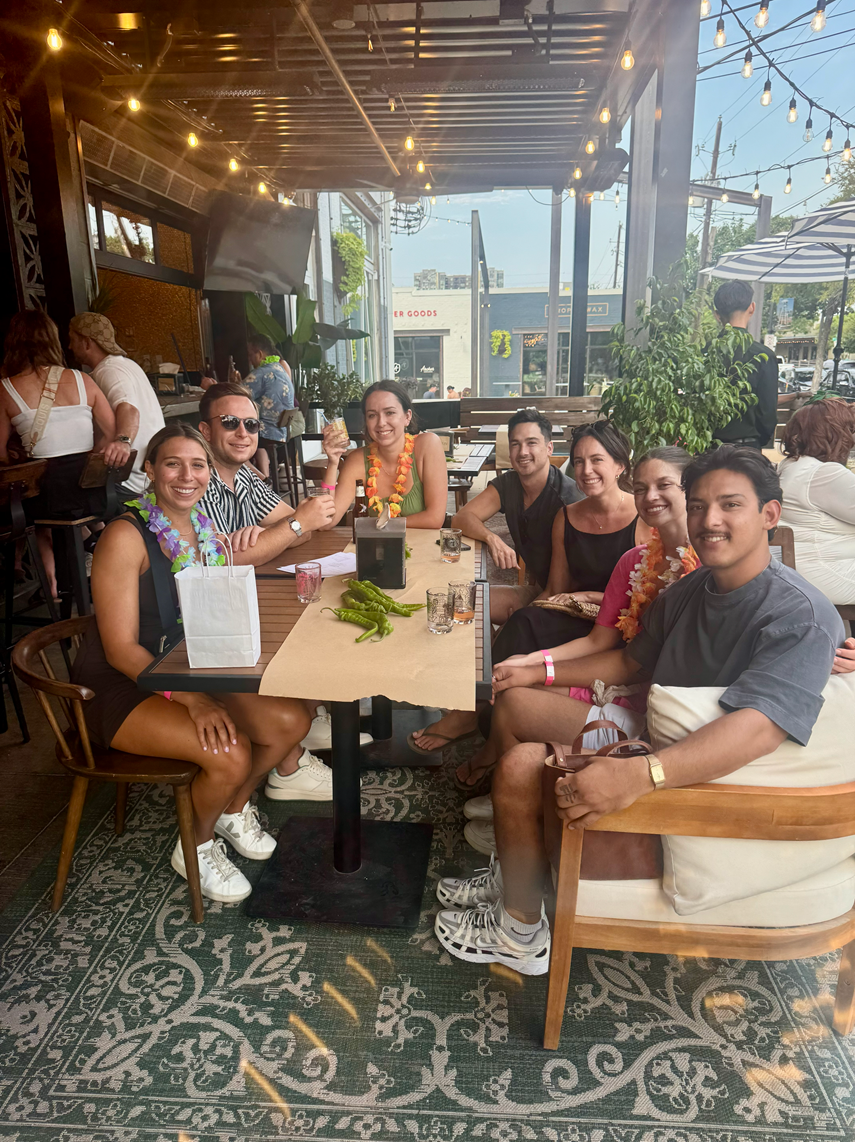 Group of seven friends sitting around a table at a restaurant enjoying drinks, with some wearing leis and a casual indoor-outdoor setting with string lights and outdoor scenery in the background.