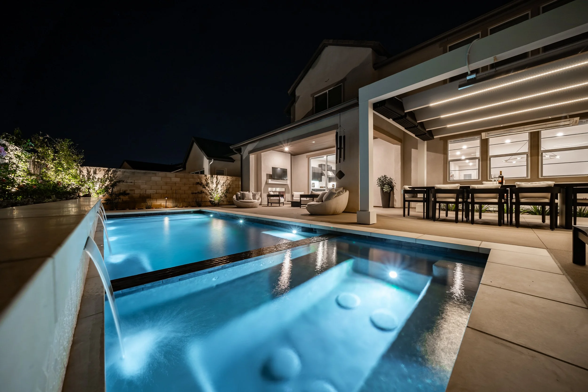 Nighttime view of a backyard swimming pool with underwater lights, surrounded by outdoor furniture under a covered patio with modern lighting, and a house in the background.