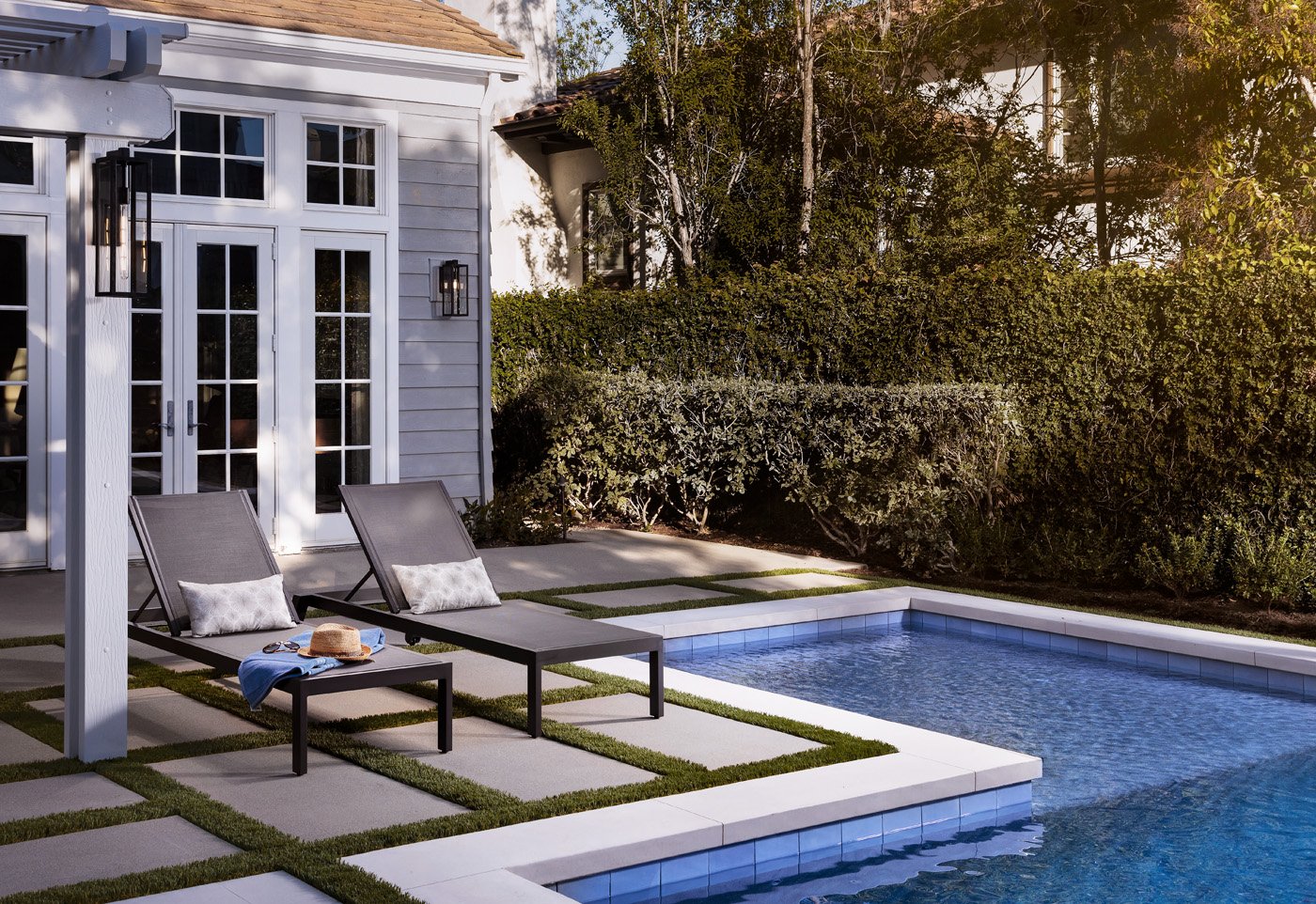 Backyard with swimming pool, two lounge chairs with pillows, a small table with a hat and towel, surrounded by greenery and a white house with glass door and outdoor lights.