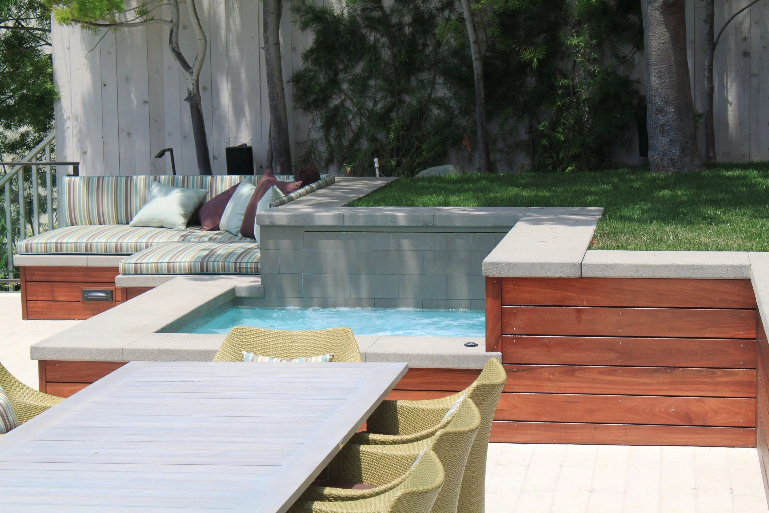 A backyard patio with a striped outdoor sofa, a wooden dining table with chairs, a swimming pool, a grassy area, and trees with a wooden fence in the background.
