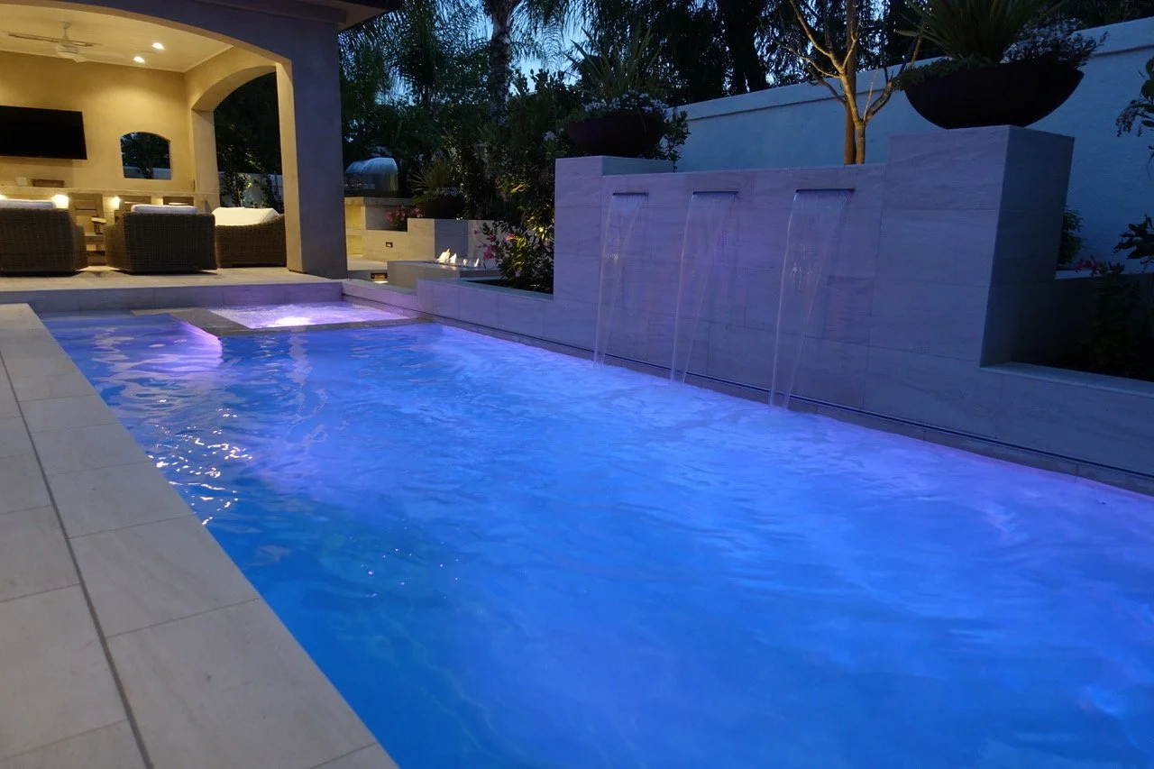 A backyard swimming pool illuminated with blue and purple lights, featuring a water wall with multiple streams, an outdoor seating area with wicker chairs and a television, and lush greenery surrounding the space during dusk.