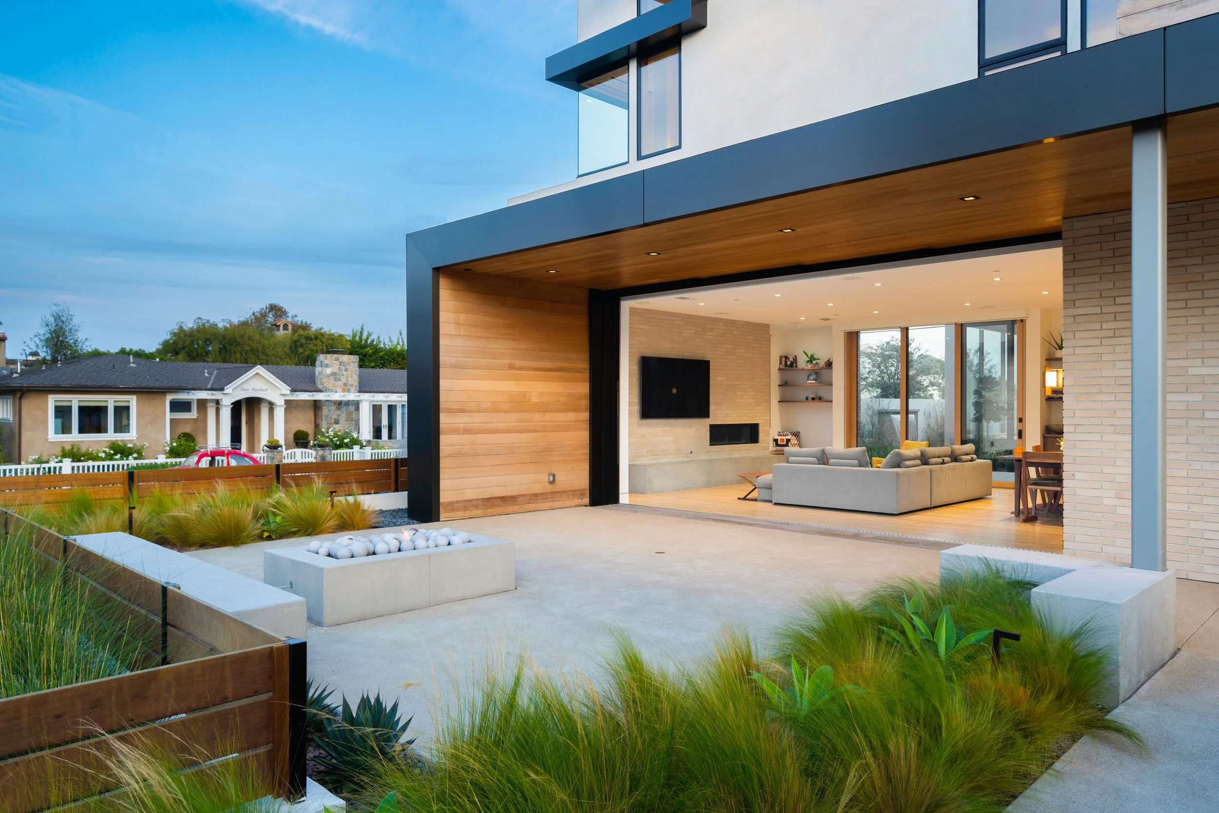 Modern house with an open living room visible through large sliding glass doors, outdoor patio with white decorative stones, wooden ceiling accents, and landscaped yard with green plants, neighboring houses in background, and a partly cloudy sky.