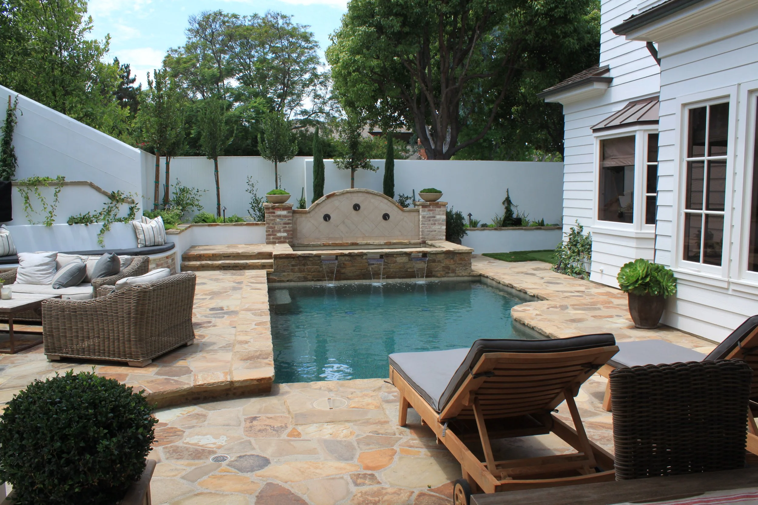 Backyard patio with a swimming pool, outdoor seating, potted plants, a water fountain, and a white house with windows, surrounded by trees and a white fence.