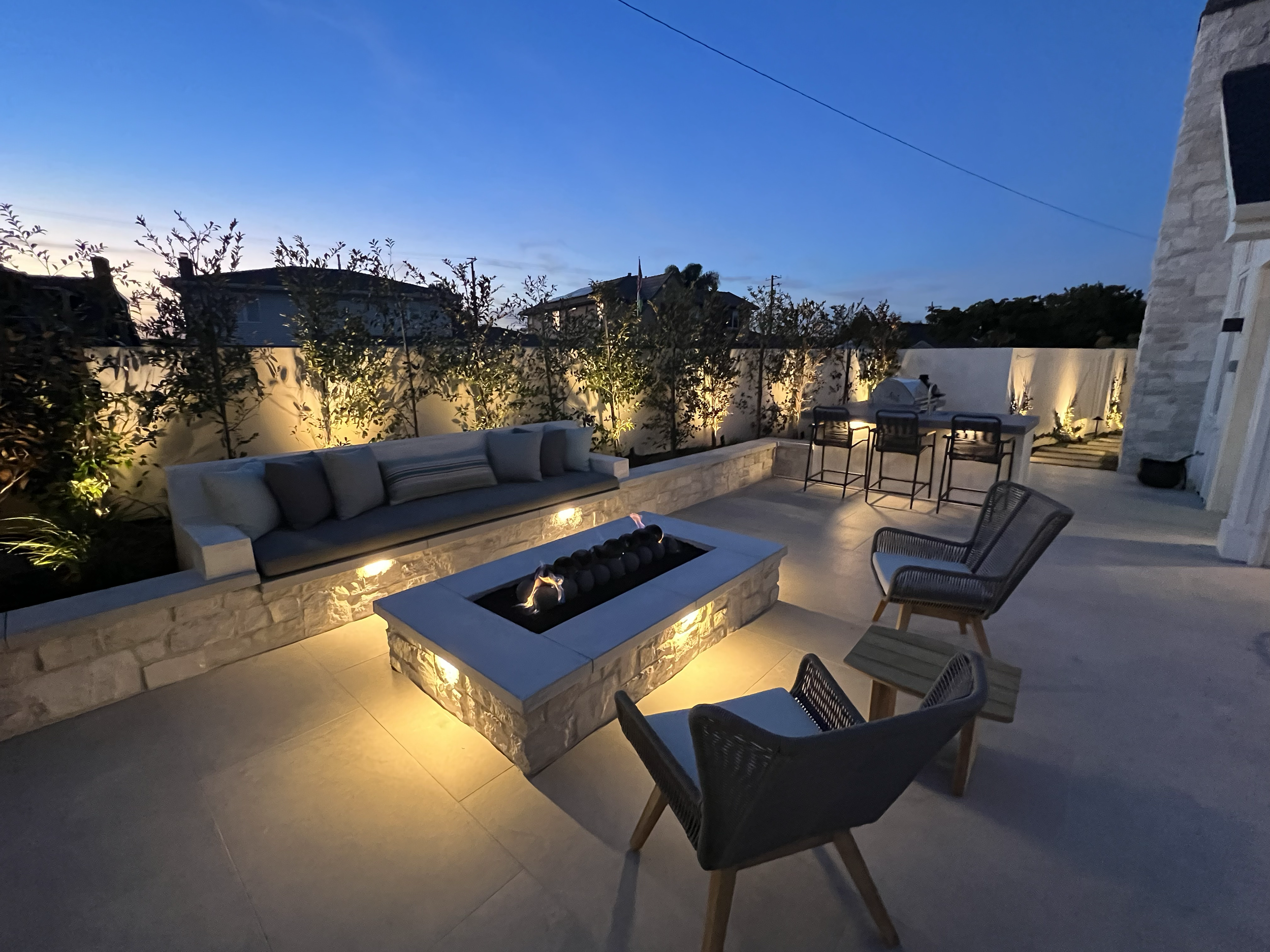 Outdoor patio area at dusk featuring a concrete fire pit, cushioned seating, dining table with chairs, and decorative lighting along a white privacy fence with trees behind it.