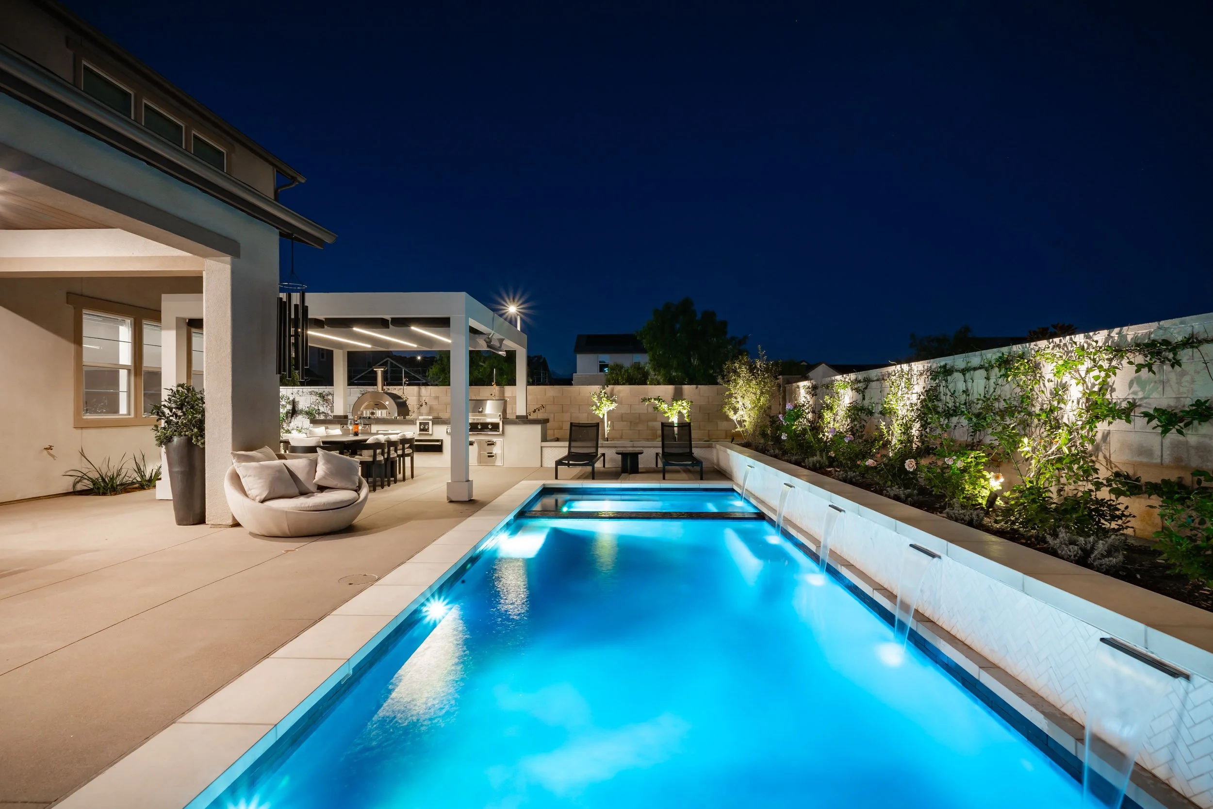 Nighttime view of a modern backyard with a lit swimming pool, outdoor seating, a built-in grill, and string lights, enclosed by a privacy wall with greenery.