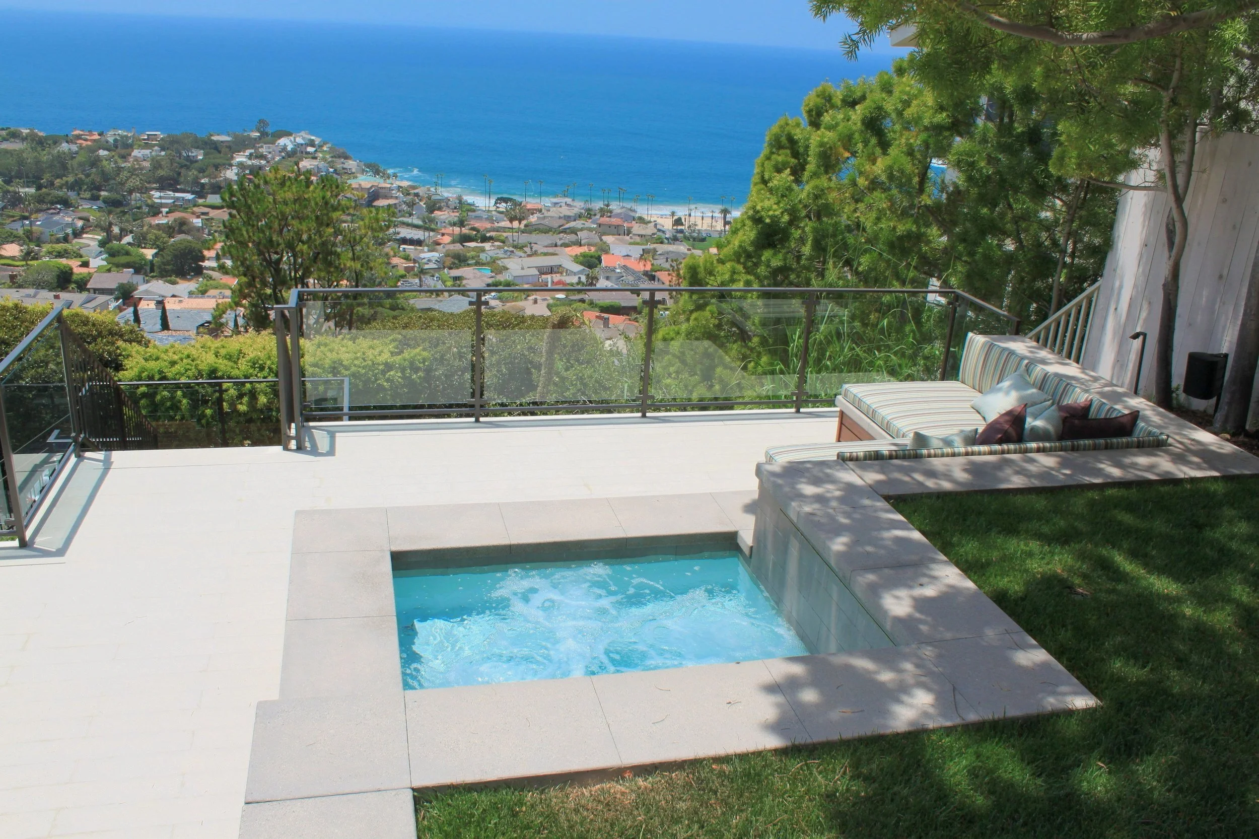 A backyard view with a swimming pool, lounge seating with striped cushions, and a scenic ocean and coastal neighborhood in the background. There are trees and greenery around the patio area.