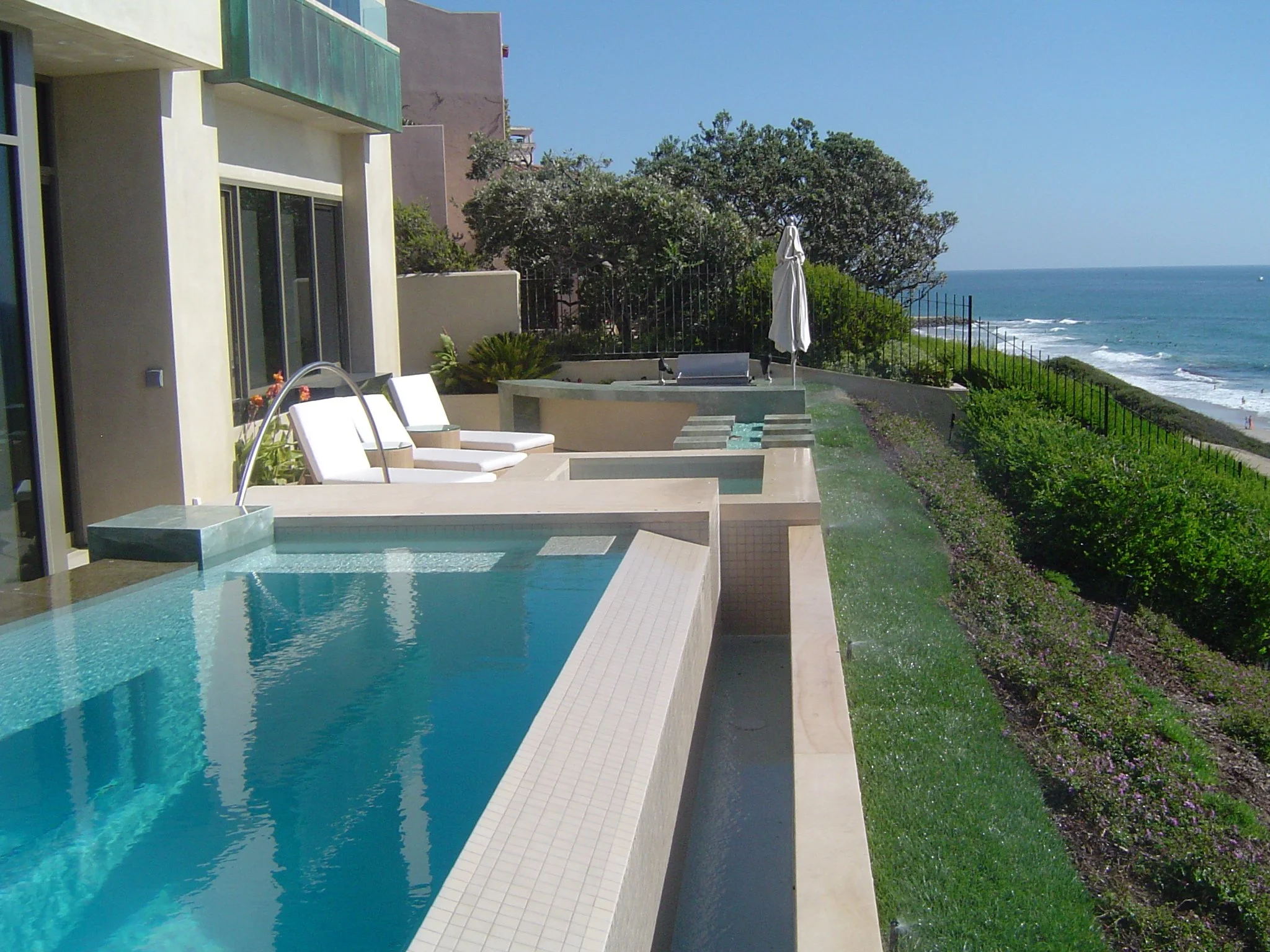 Modern house balcony with a pool, lounge chairs, and an ocean view, including trees, an umbrella, and a fence along the coastline.