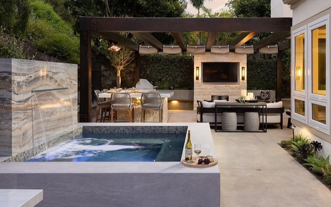 Outdoor patio with hot tub, dining area, and lounge with a large flat-screen TV, surrounded by greenery and modern decor.