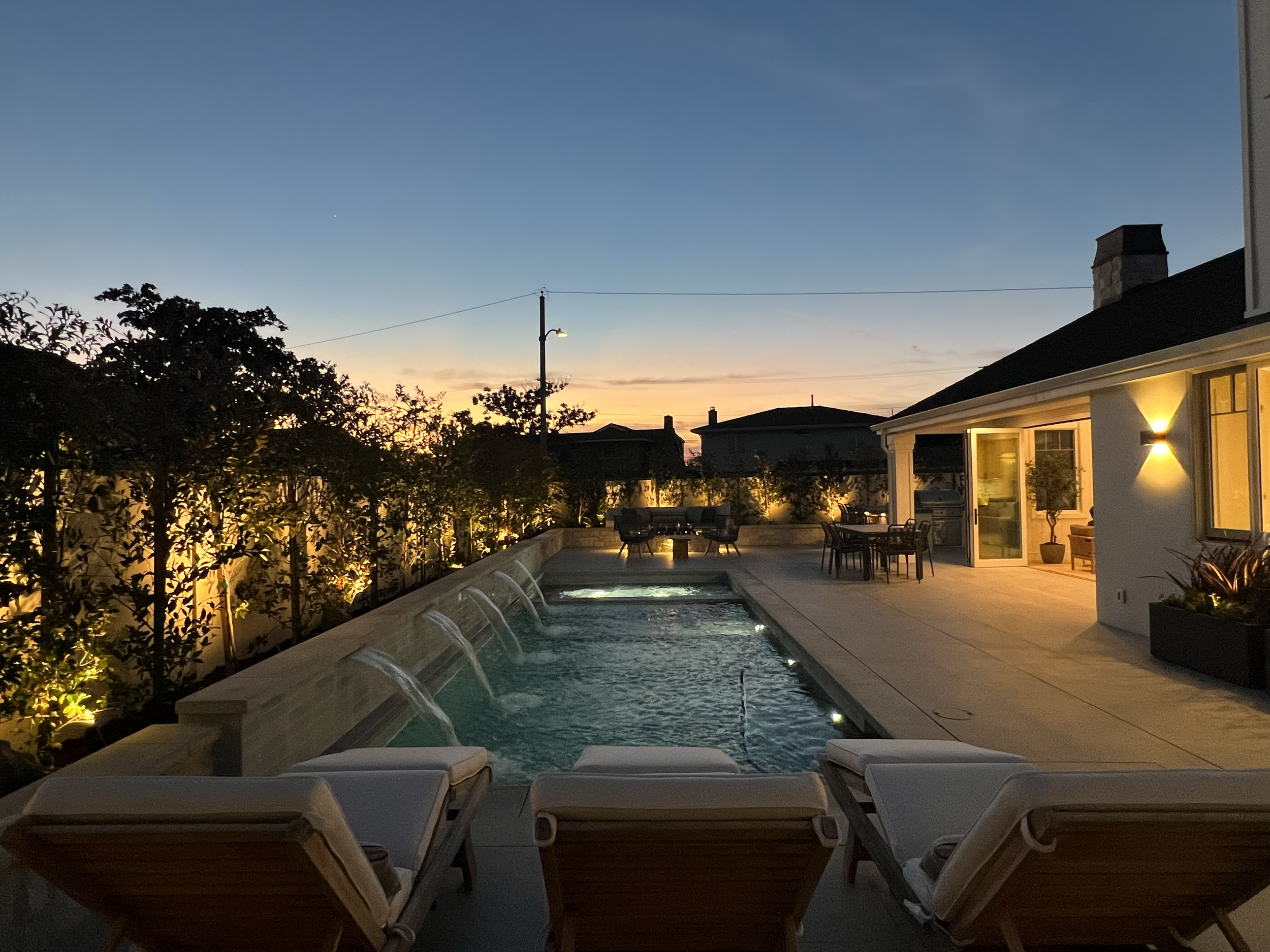 A backyard pool area at dusk with lounge chairs, a screened porch with outdoor furniture, trees, and a sunset sky with faint clouds overhead.