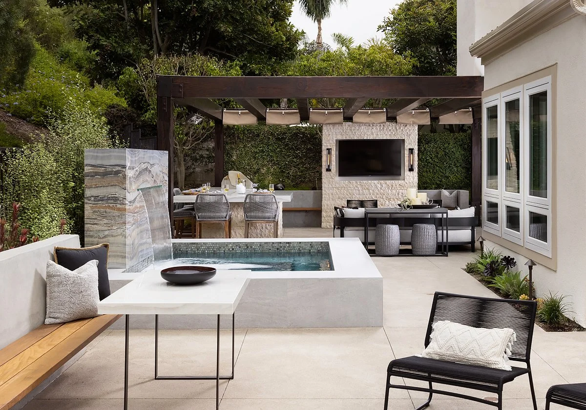 Outdoor patio featuring a small pool, a bench with pillows, a table, a seating area with a TV, a dining table with chairs, and lush greenery in the background.
