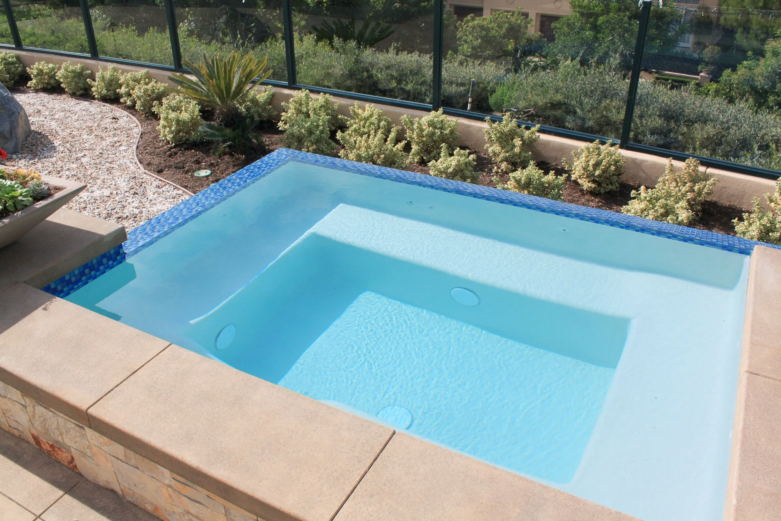 Round, built-in hot tub or swim spa with blue tile border, situated outdoors on a paved patio, surrounded by landscaped garden beds and a glass fence, with trees and a building visible in the background.