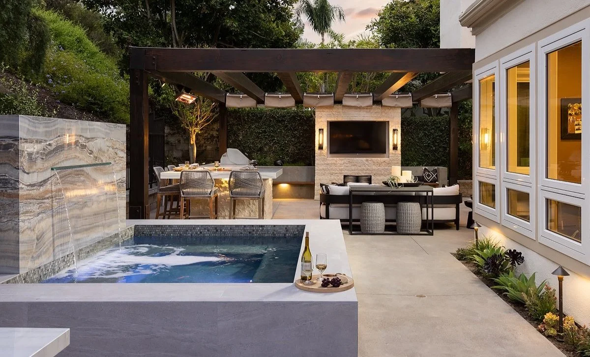 Modern backyard with a hot tub, outdoor dining area, and a seating area with a flat-screen TV mounted on stone wall, surrounded by greenery and outdoor lighting.