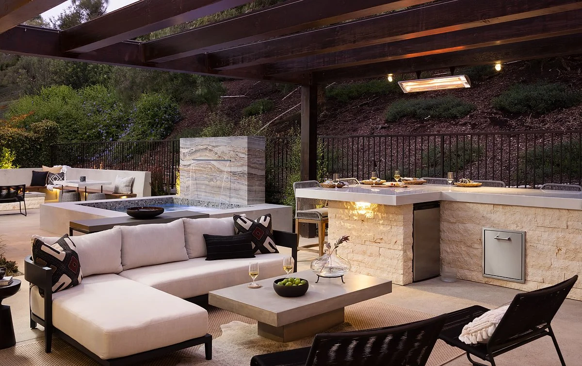 Outdoor patio with seating area, modern fireplace, bar counter, and scenic hillside background.