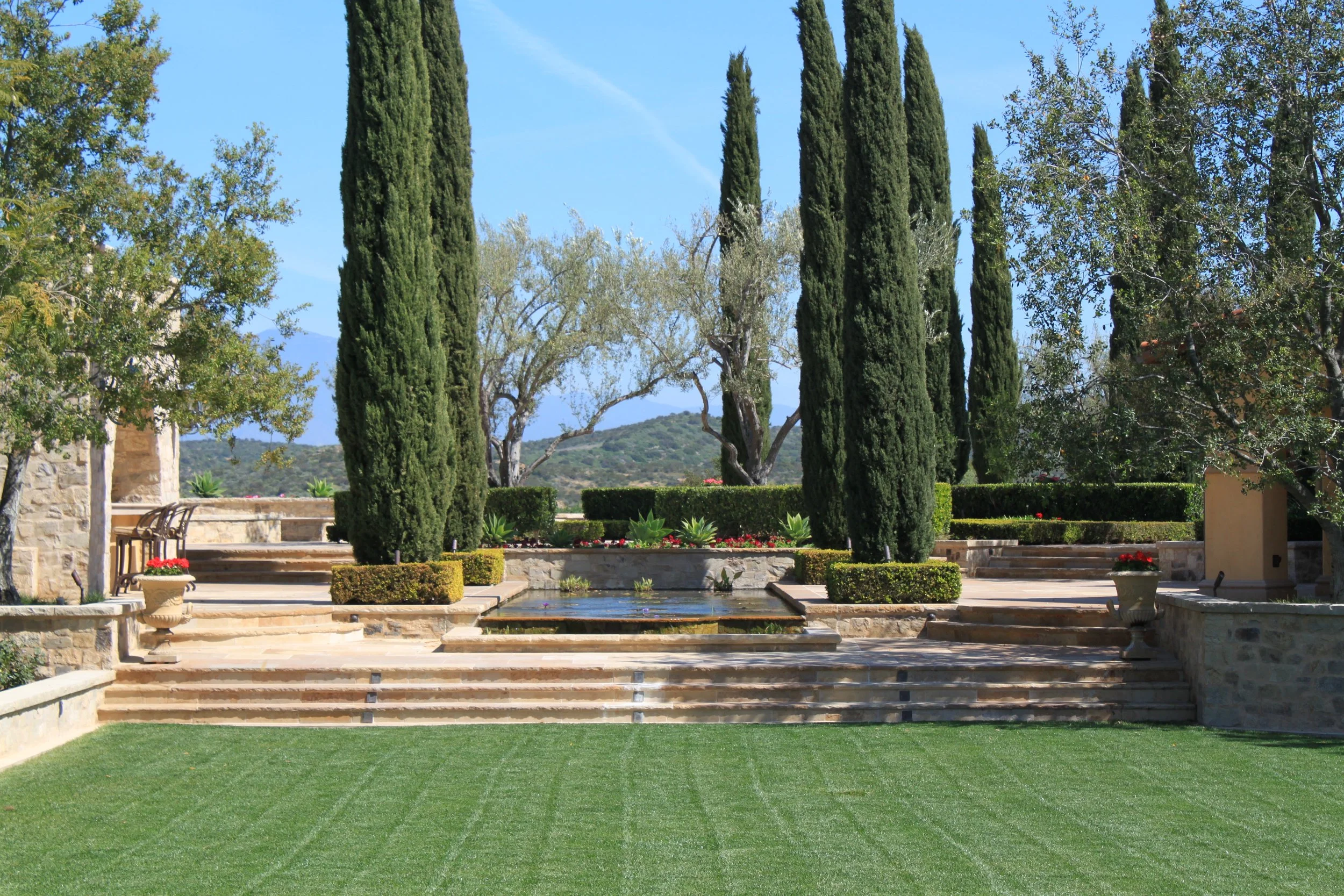 A landscaped garden with neatly trimmed grass, stone steps, and a rectangular water feature. Tall cypress trees and other greenery surround the area, with a stone building partially visible on the left. Clear blue sky overhead.