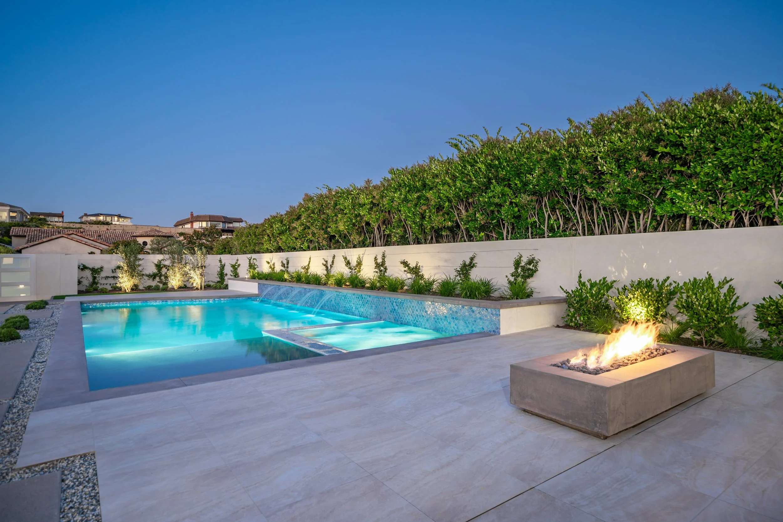 Modern backyard with illuminated swimming pool, fire feature, lush green hedge, and neighboring houses under a clear blue sky.