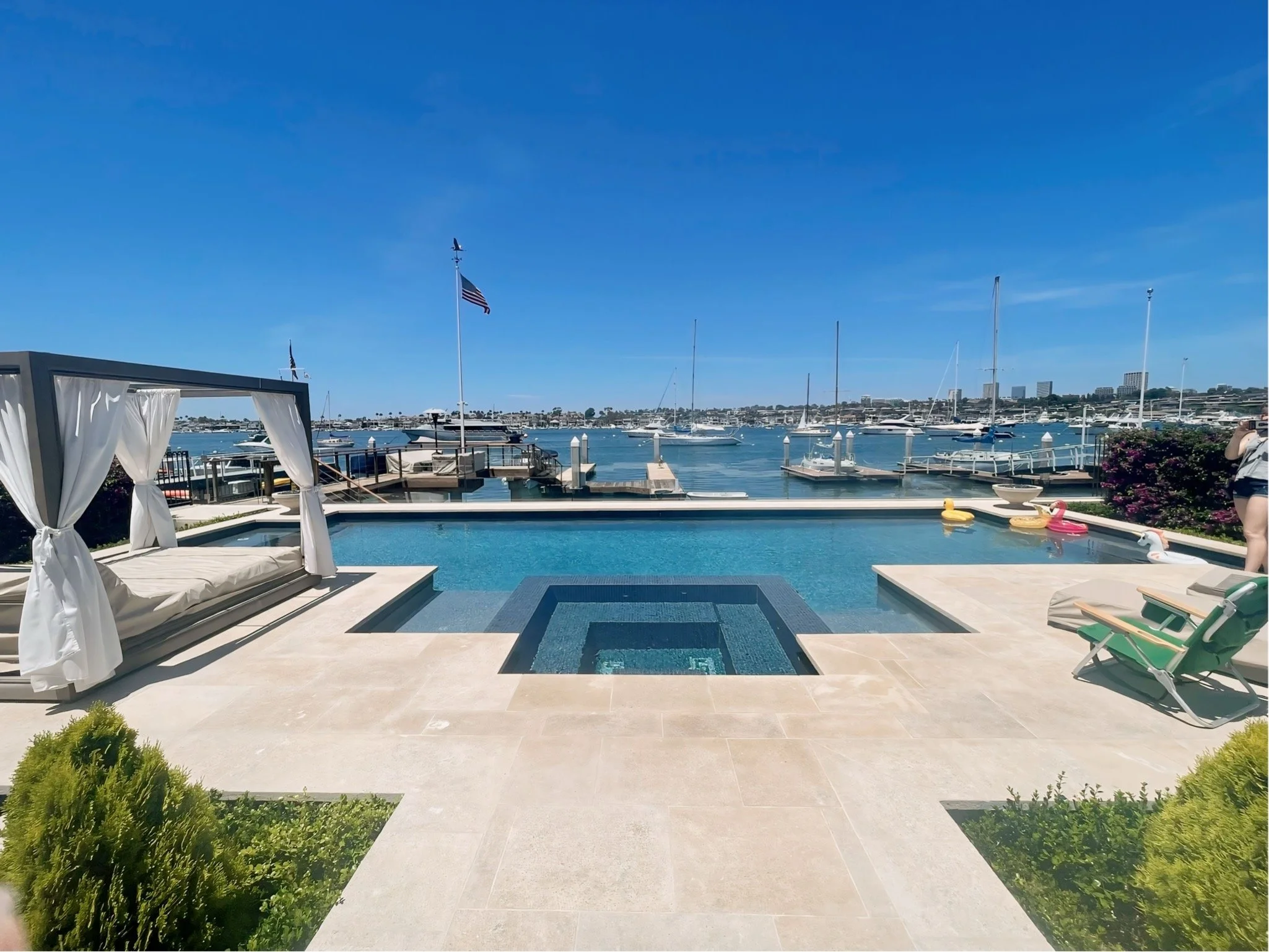A view of a marina with boats docked on calm water, surrounded by a clear blue sky. In the foreground, there is a swimming pool with pool floaties shaped like animals and a lounge chair with white curtains. Green bushes are on the edges.