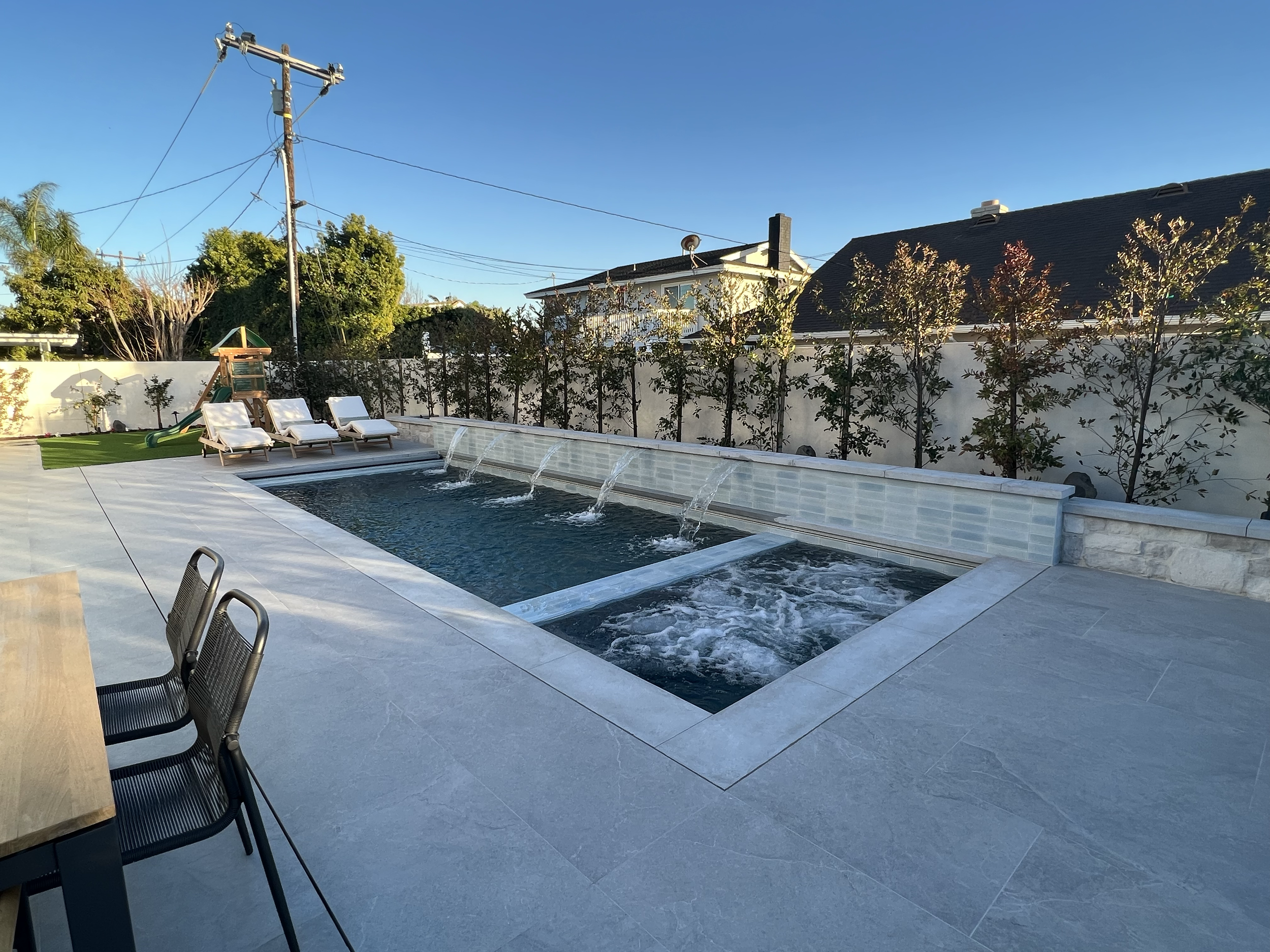 A backyard pool area with three white lounge chairs, a small slide, and a landscaped border with trees and shrubs, under a clear blue sky.