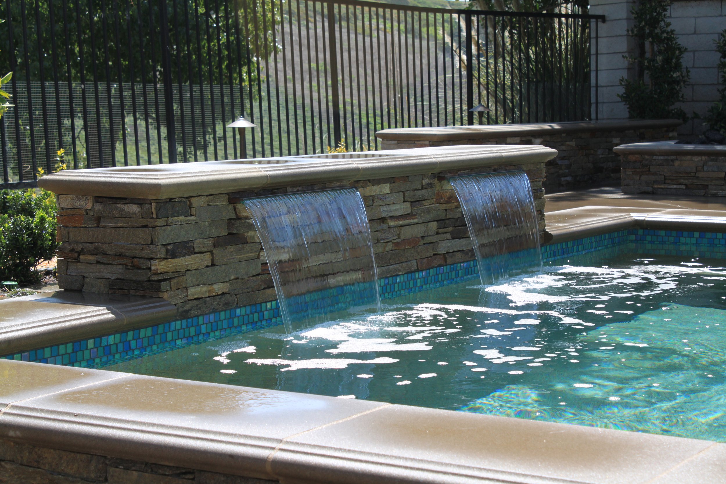 A backyard pool features a stone waterfall with water cascading into the pool, surrounded by a stone deck and a black metal fence with garden lights, trees, and a brick wall in the background.