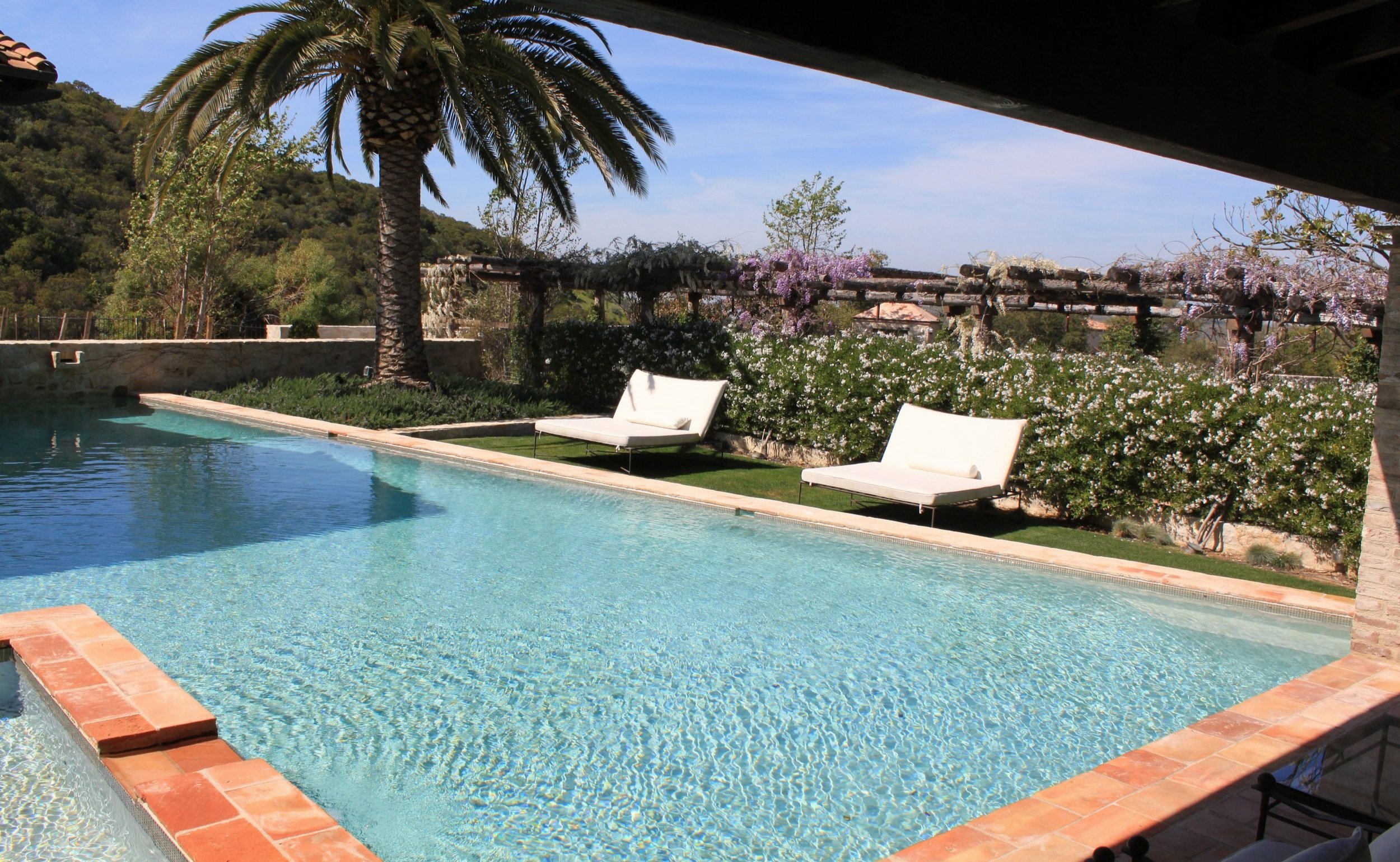 A backyard swimming pool with clear blue water, surrounded by a brick edge, featuring two white lounge chairs on the grass nearby. In the background, there are lush green trees, flowering bushes with white and purple blossoms, and a hillside with den