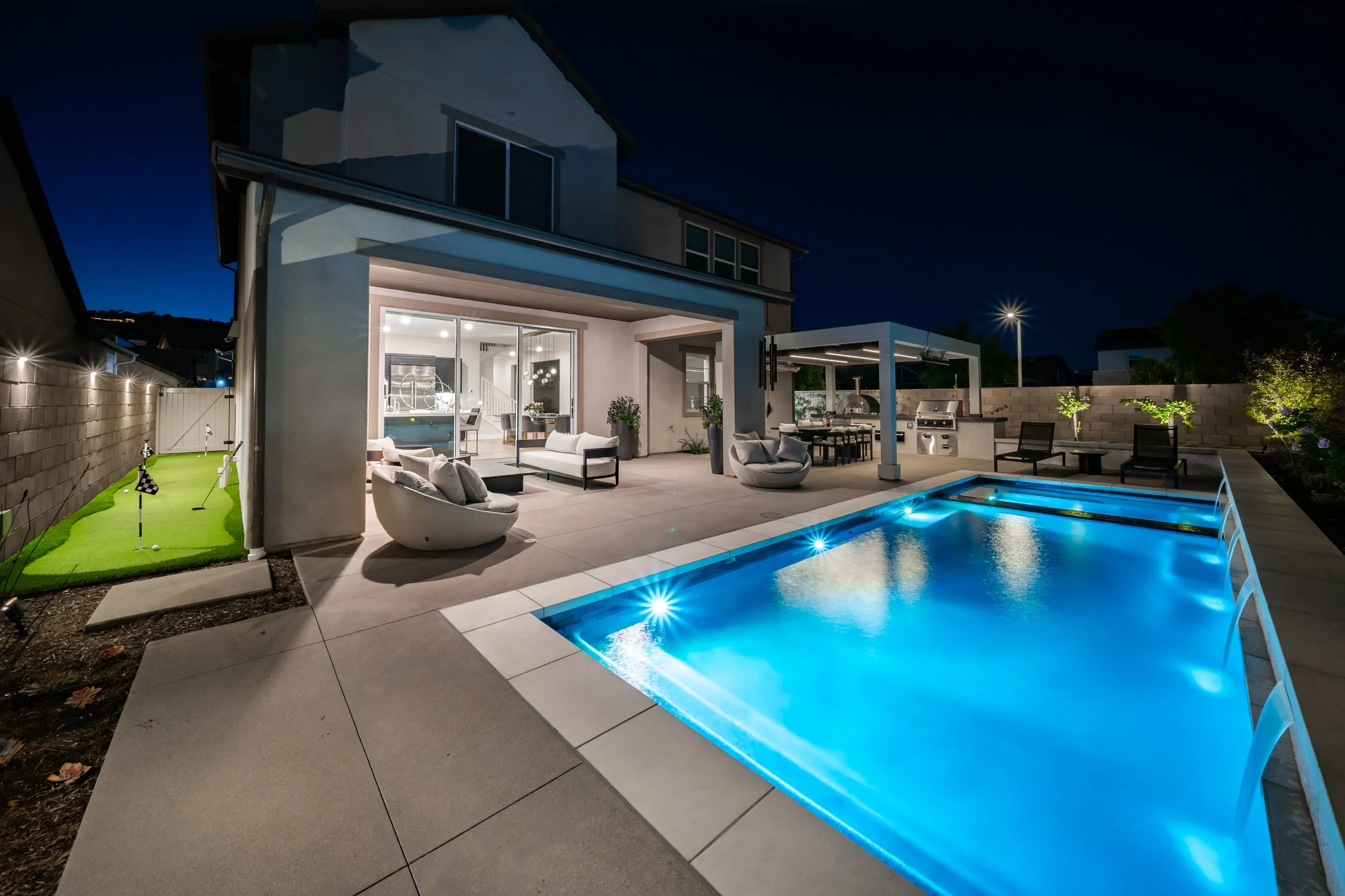 A modern backyard at night with a lit swimming pool, outdoor seating, a grill, and a mini-golf putting green against a fence, with a house featuring large glass doors and windows illuminated from inside.