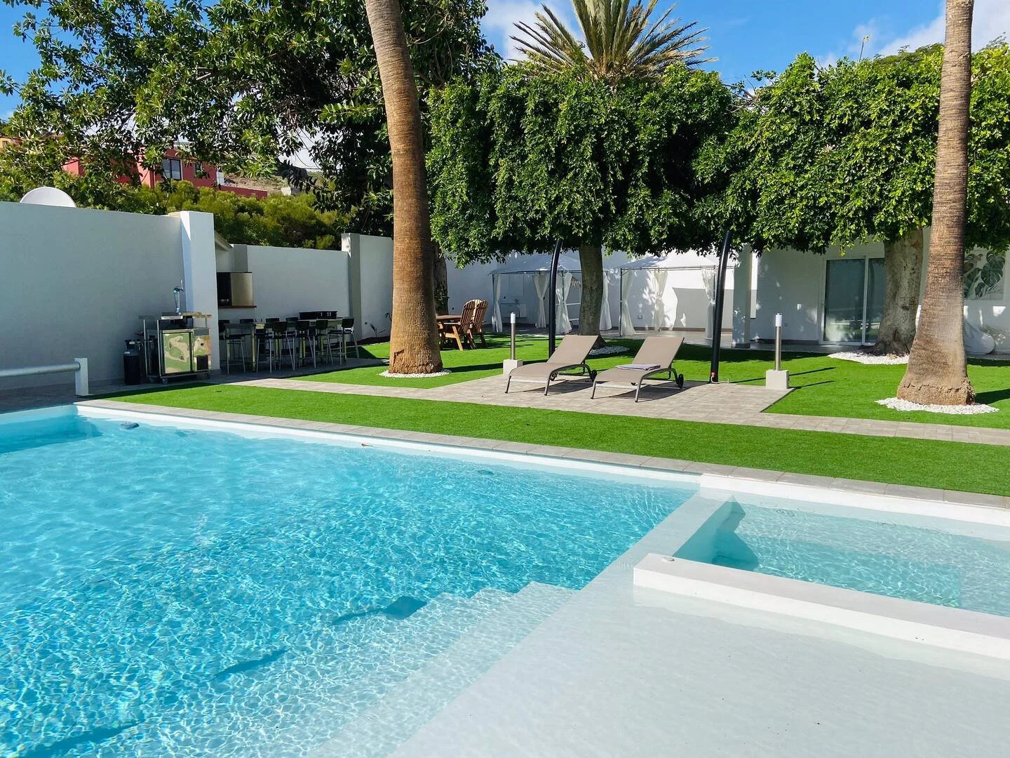 Luxury backyard with a swimming pool, two lounge chairs under a shaded tree, a grassy area with a paved walkway, a bar area with stools, and outdoor dining chairs.