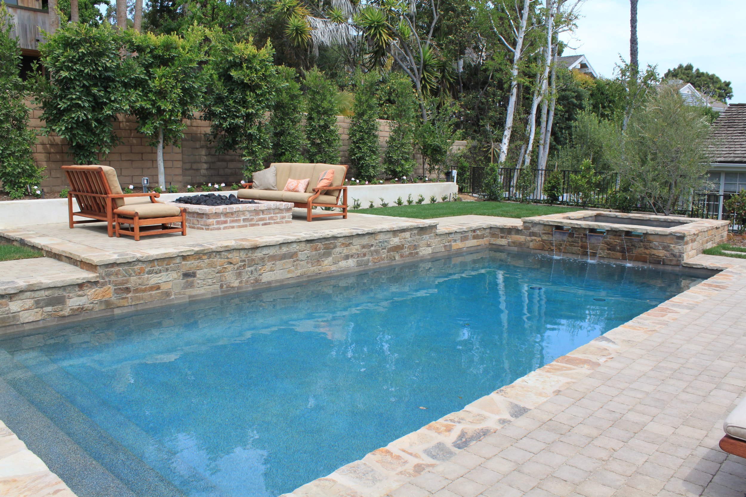 backyard scene with a swimming pool, outdoor furniture including a cushioned loveseat and chairs, a fire pit, lush green trees, and a brick wall in the background.