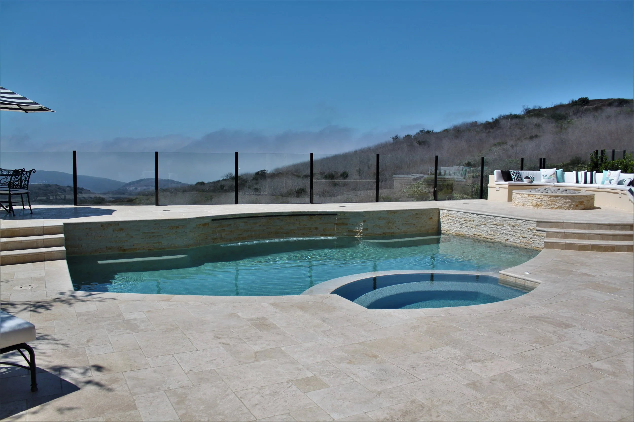 A swimming pool with a curved edge, surrounded by a stone deck. There are built-in lounge chairs with cushions on the right side. In the background, there is a glass fence and scenic hills under a clear blue sky.
