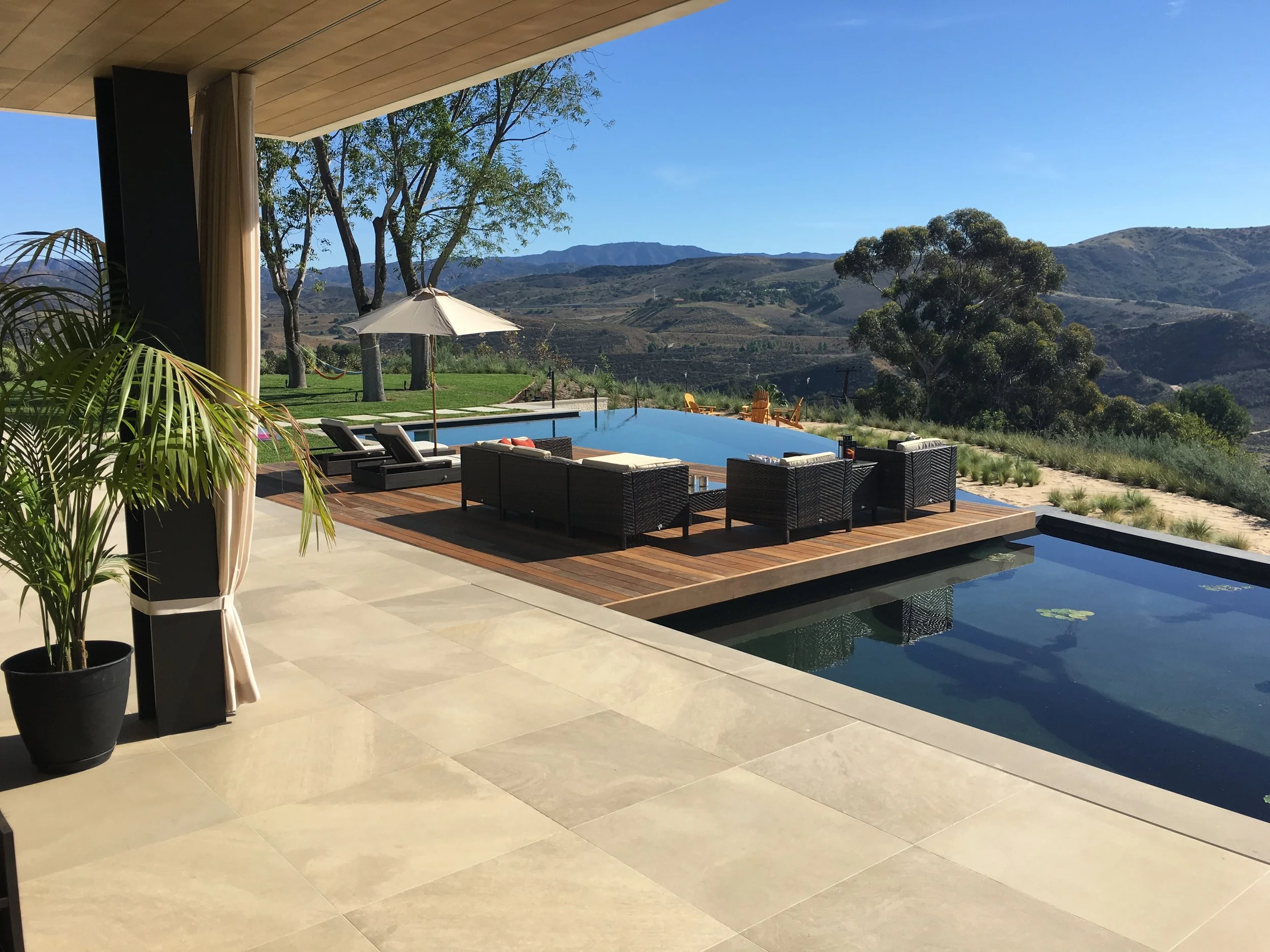 Outdoor pool area with decking, surrounded by outdoor furniture including lounge chairs and an umbrella, with a scenic mountain view in the background under a clear blue sky.