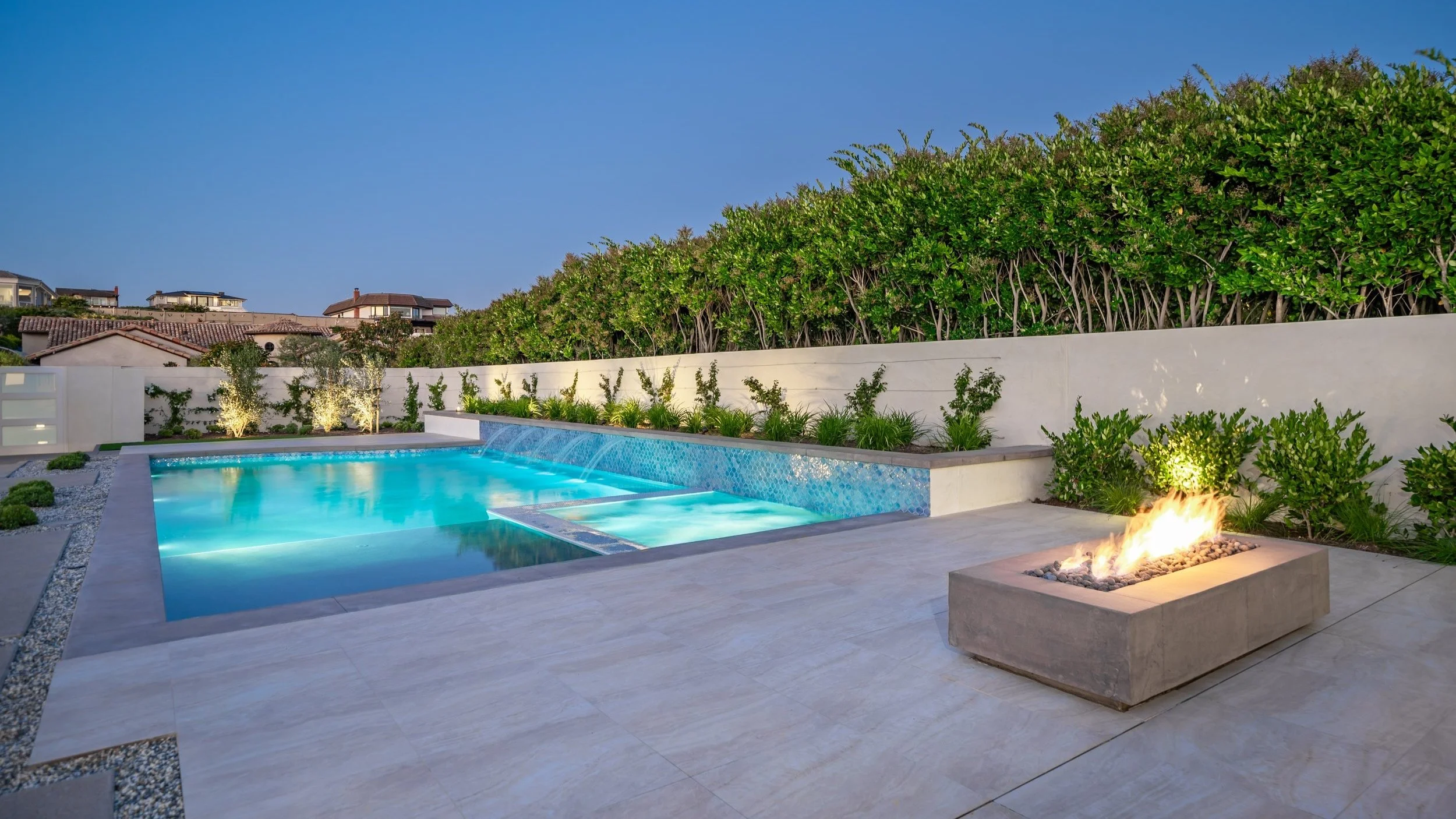 Modern backyard with a swimming pool, fire feature, and lush green hedge wall during twilight.