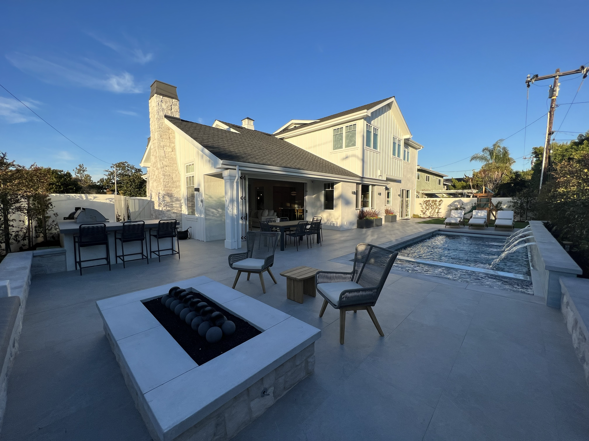 Backyard with a swimming pool, outdoor seating, and a large white house with multiple windows. There is a fire pit with black stones and a wooden dining table with chairs near the house. The sky is clear with few clouds.