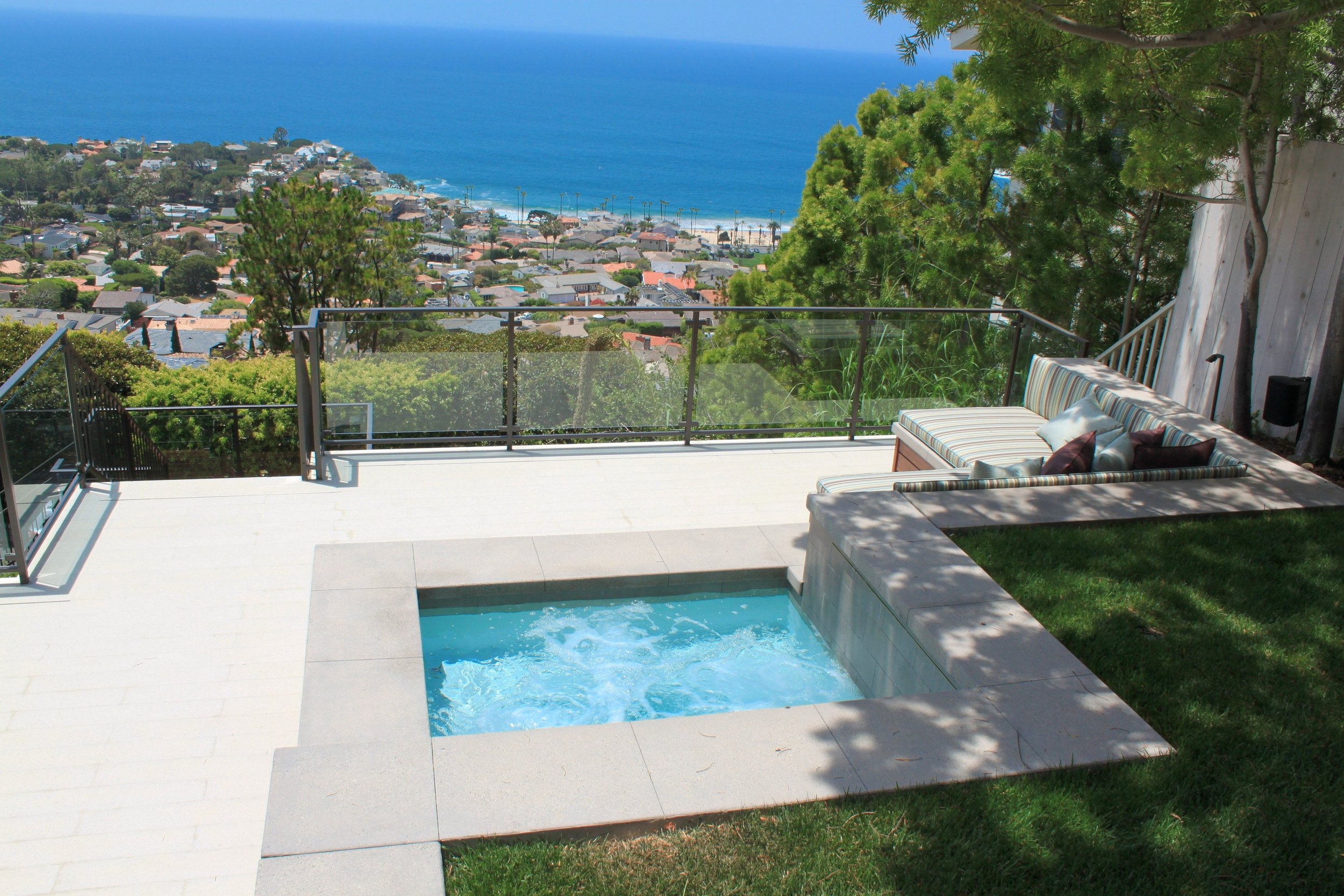 A scenic view from a backyard deck overlooking a neighborhood, the beach, and the ocean in the distance. The deck features a small swimming pool, a striped outdoor sofa with pillows, and a glass railing with green trees nearby.