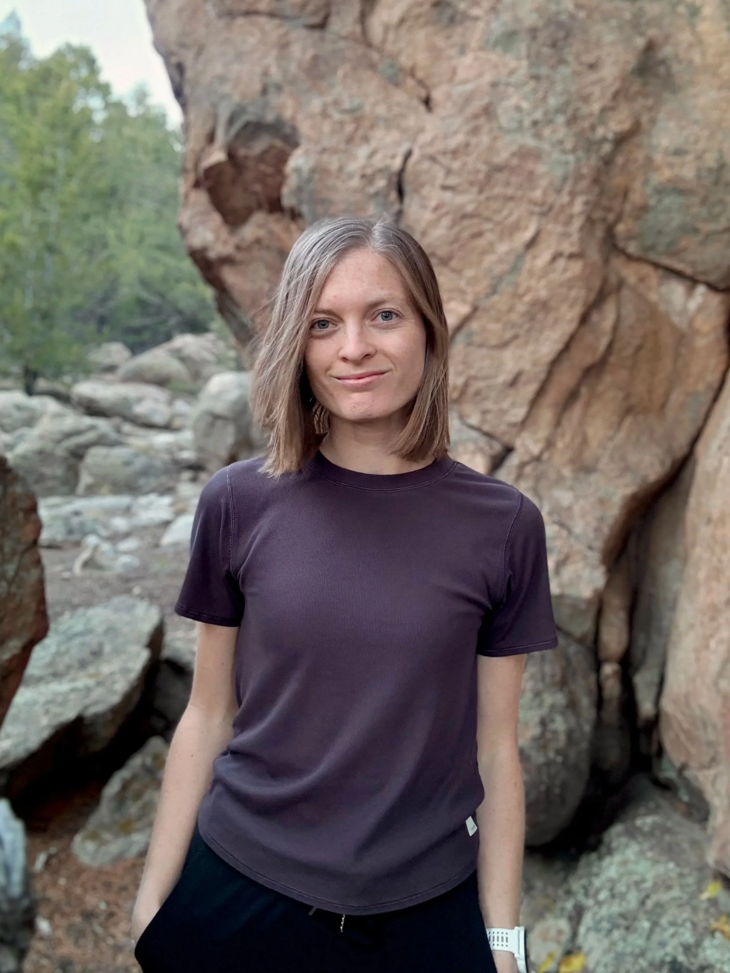 Woman stands in front of large rock with trees in the background in a natural environment in Colorado.