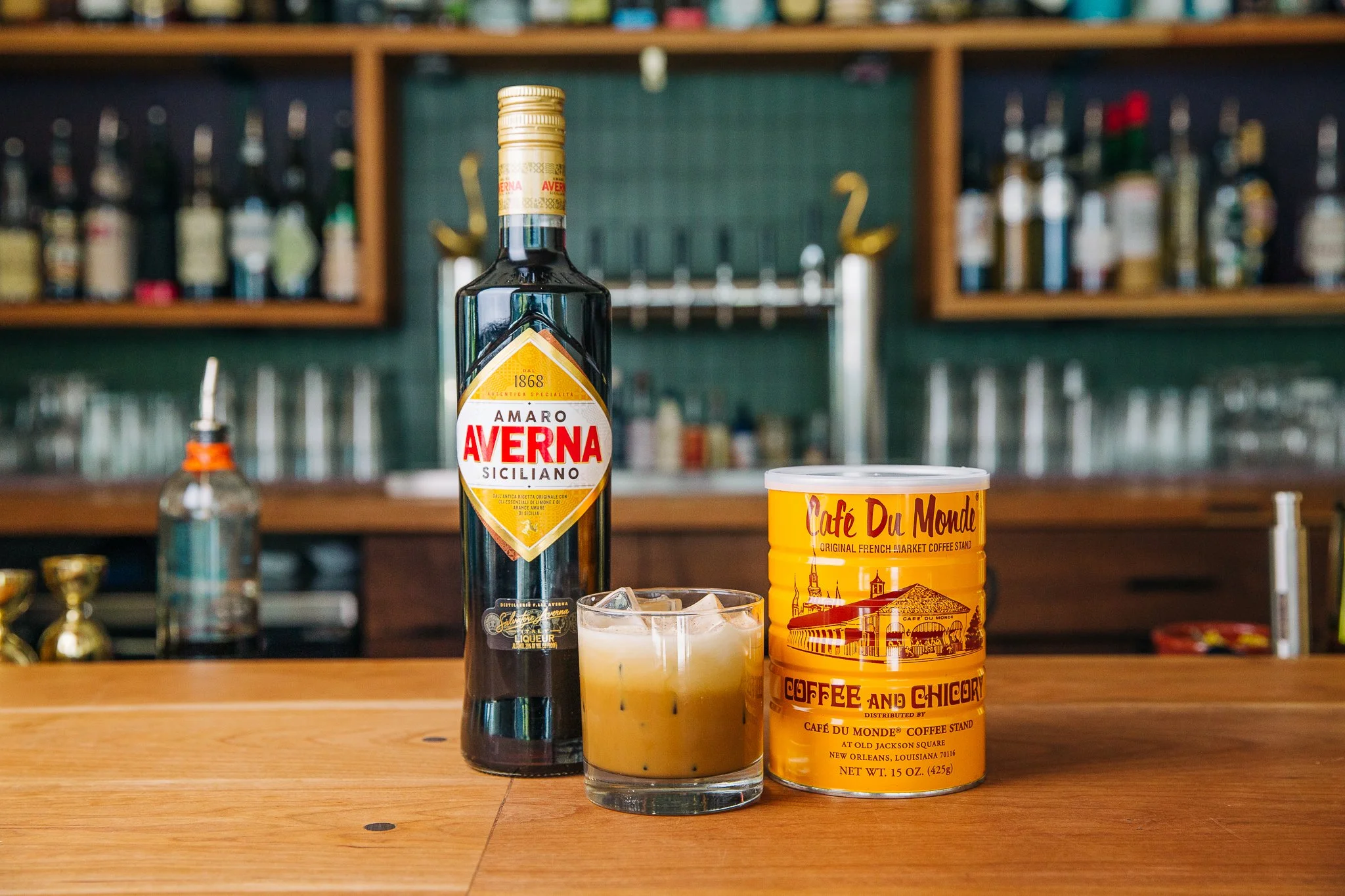 Bottle of Amaro Averna, a can of Café Du Monde coffee, and a cocktail glass with a creamy drink on a bar counter, with bottles in the backgr