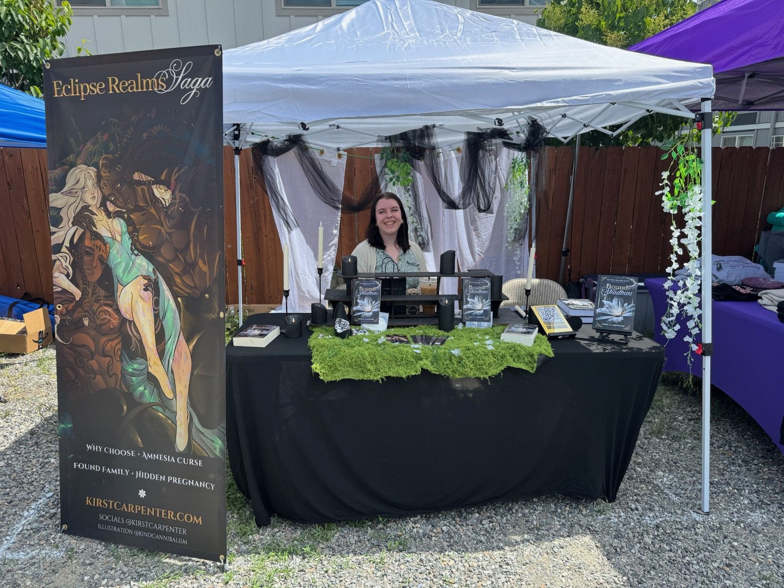 Kirst N. Carpenter sitting behind a table for a vendor event. The tablecloth is black with a green moss blanket on top, books of bound to shadows and a white canopy over the top. To the side is a banner of character art from the book.