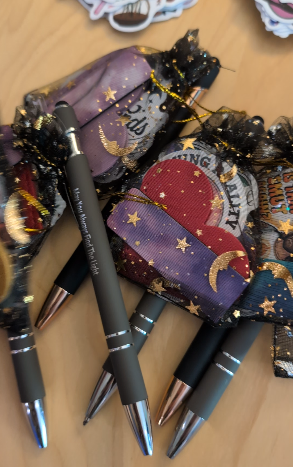 Black pens with silver tips and black bodies, placed on colorful fabric pouches featuring celestial designs. The pouches have ribbons and star patterns, one with the phrase "Find The Light." The scene appears to be on a wooden surface.