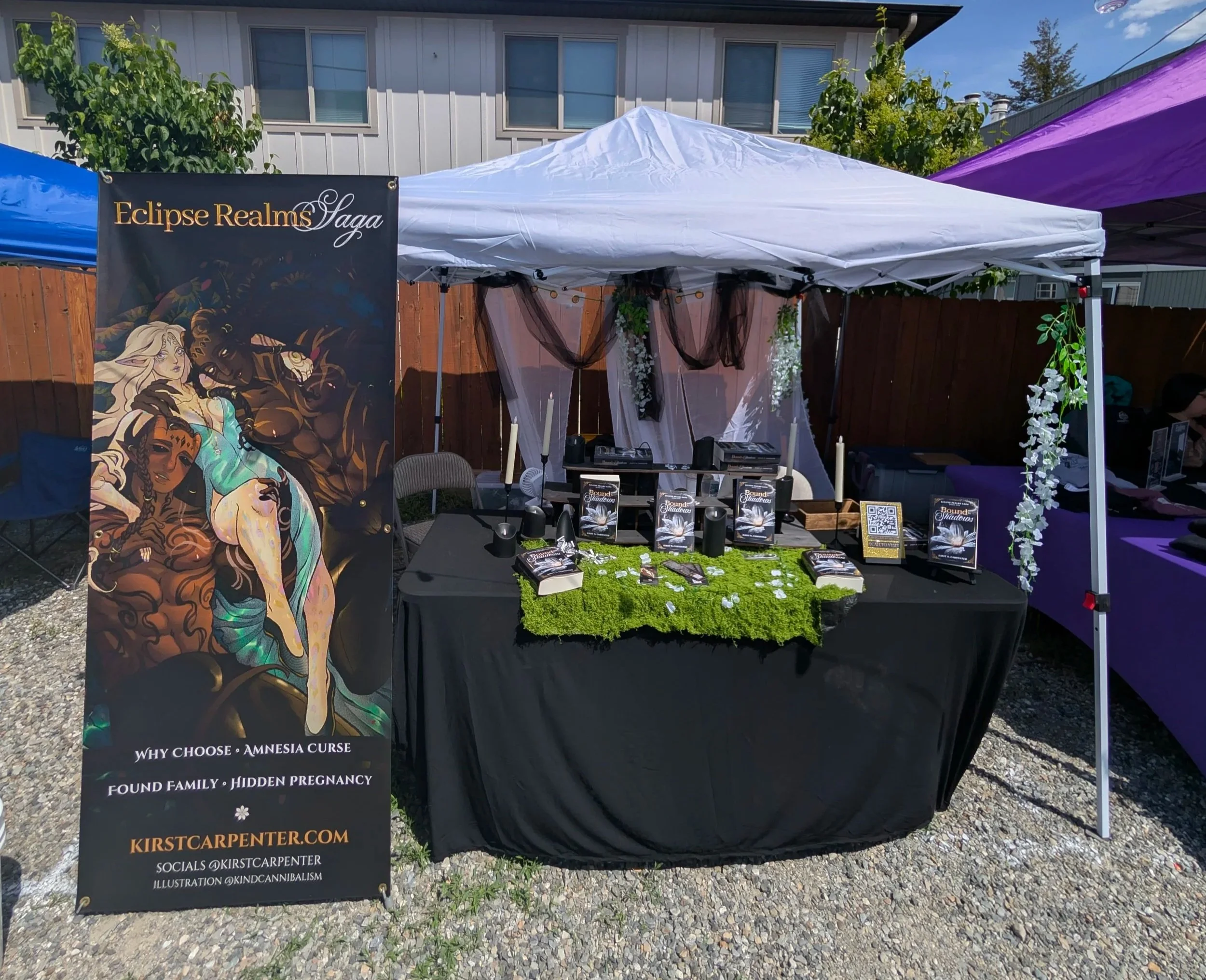 Display booth at an outdoor event with a black banner reading 'Eclipse Realms Saga' featuring fantasy art of a woman with dragons. The booth has brochures, a QR code, and a moss-covered table for promotion.